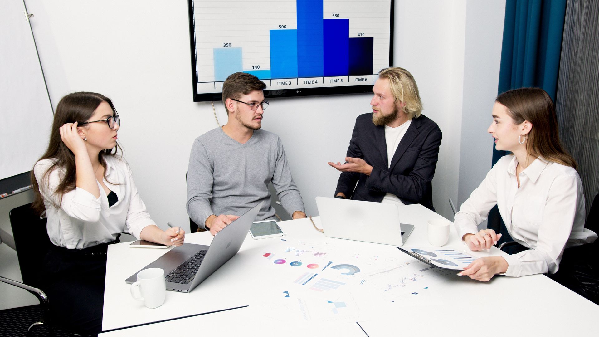 Team meeting in a bright office, four people discussing charts around a table with laptops and papers.