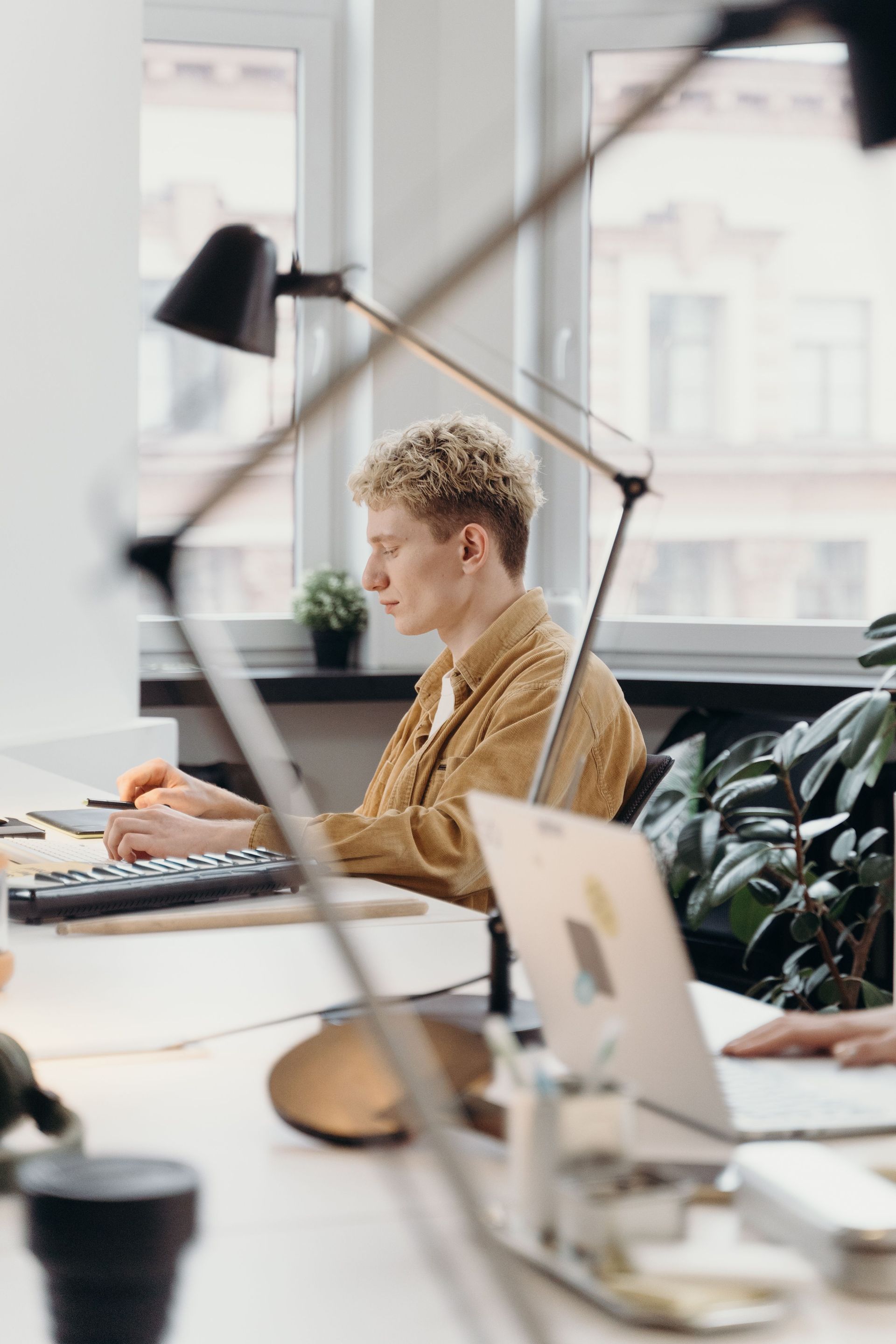 Person working at a desk in a bright office, typing on a laptop beside a lamp and coffee cup.