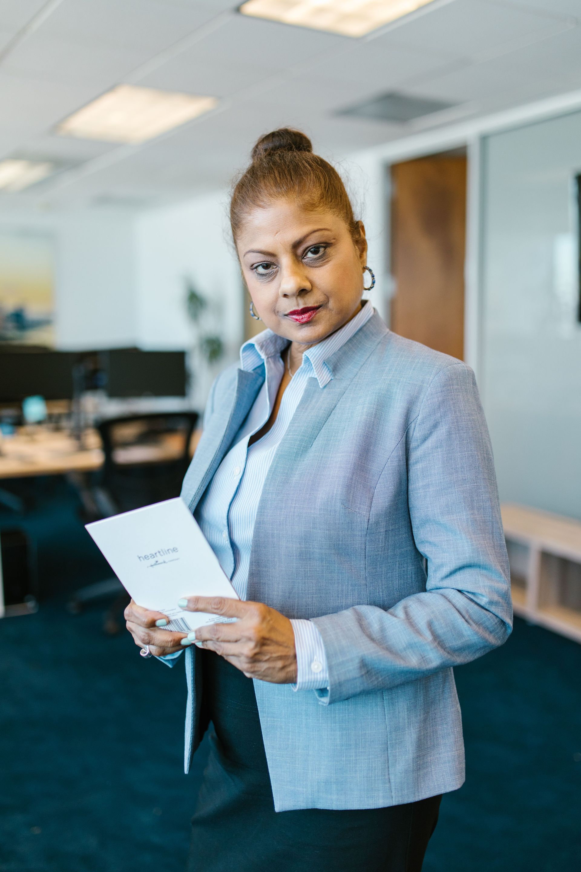 Woman in a light blue blazer holding papers in a modern office, looking at the camera.