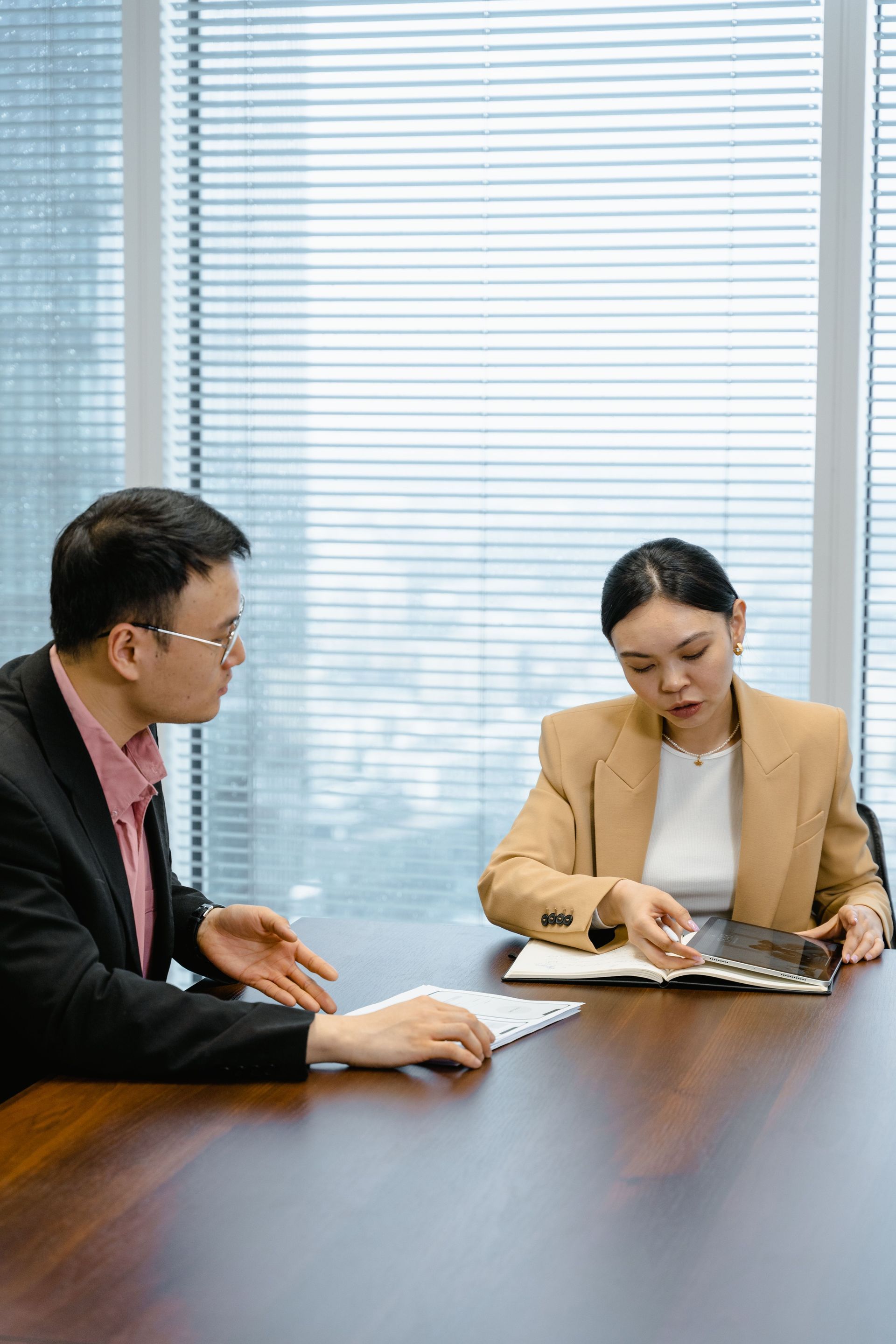 Two coworkers reviewing documents at a conference table in an office, with large blinds behind them.