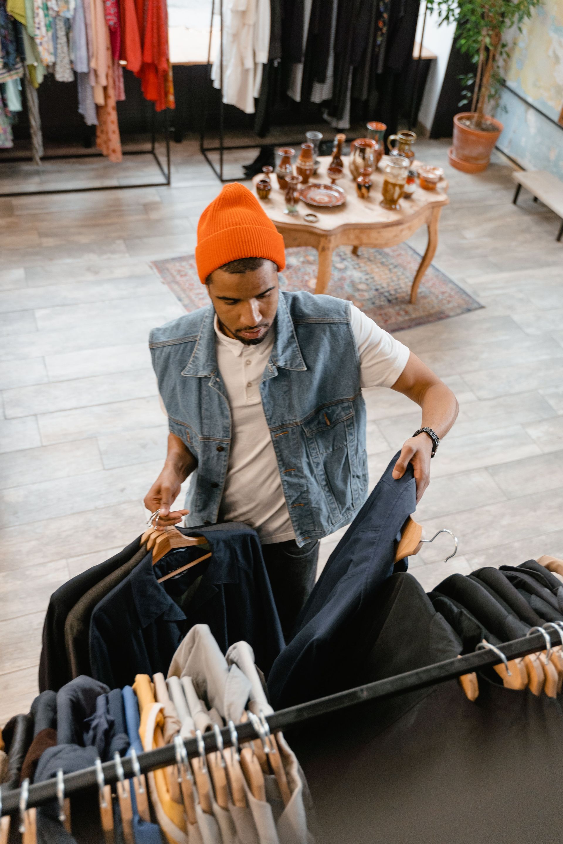 Person shopping for clothes in a boutique, holding up dark pants beside a rack of hanging garments.