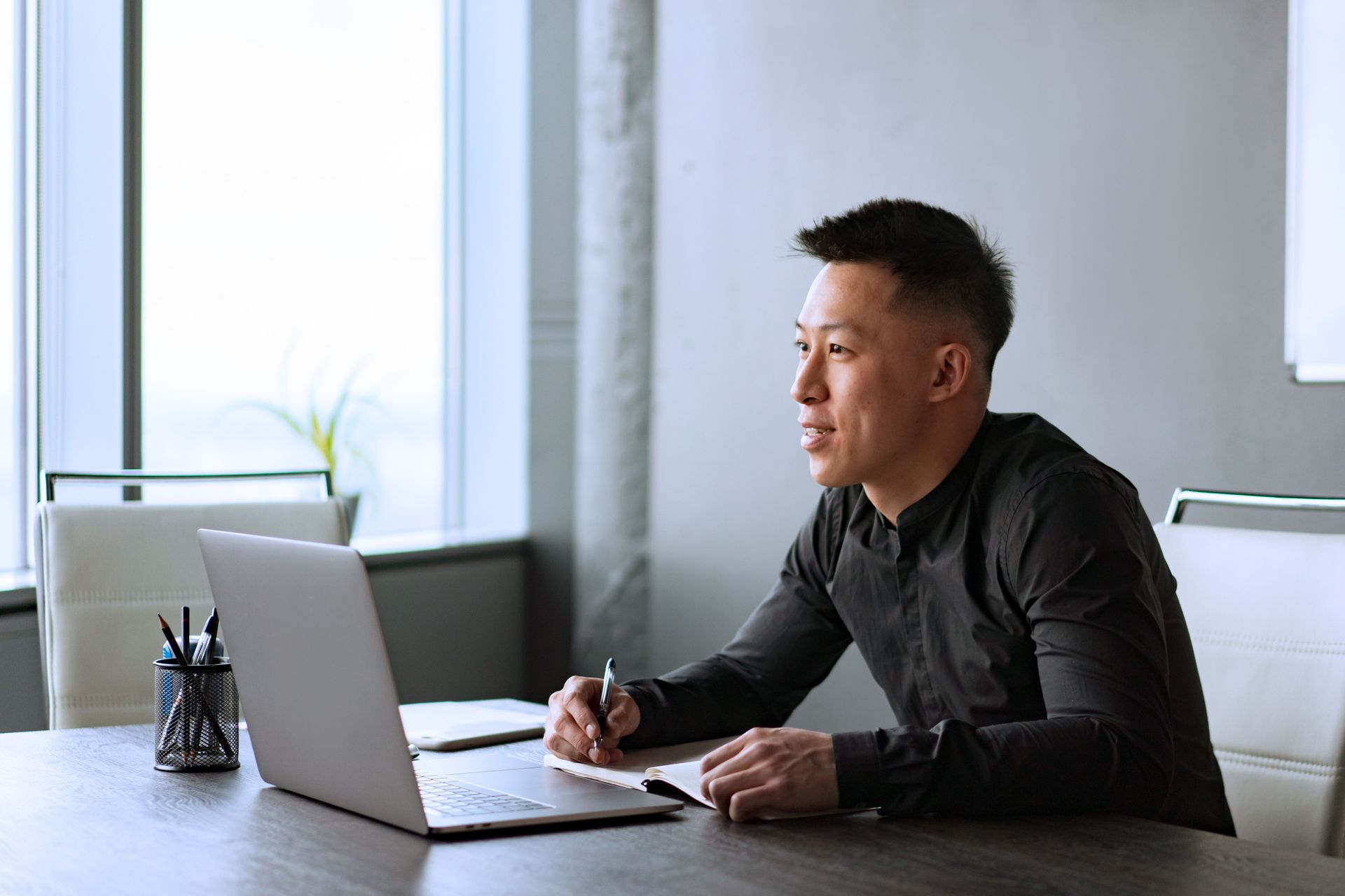 Man sitting at a desk with a laptop and notebook, writing while looking toward a window in a bright office