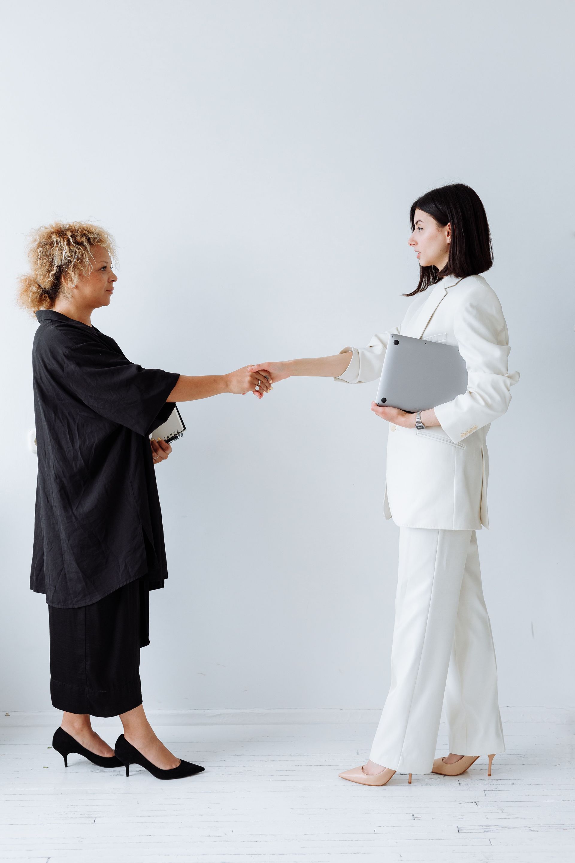 Two women shaking hands in a bright studio, one holding a clipboard and wearing white, the other in black.