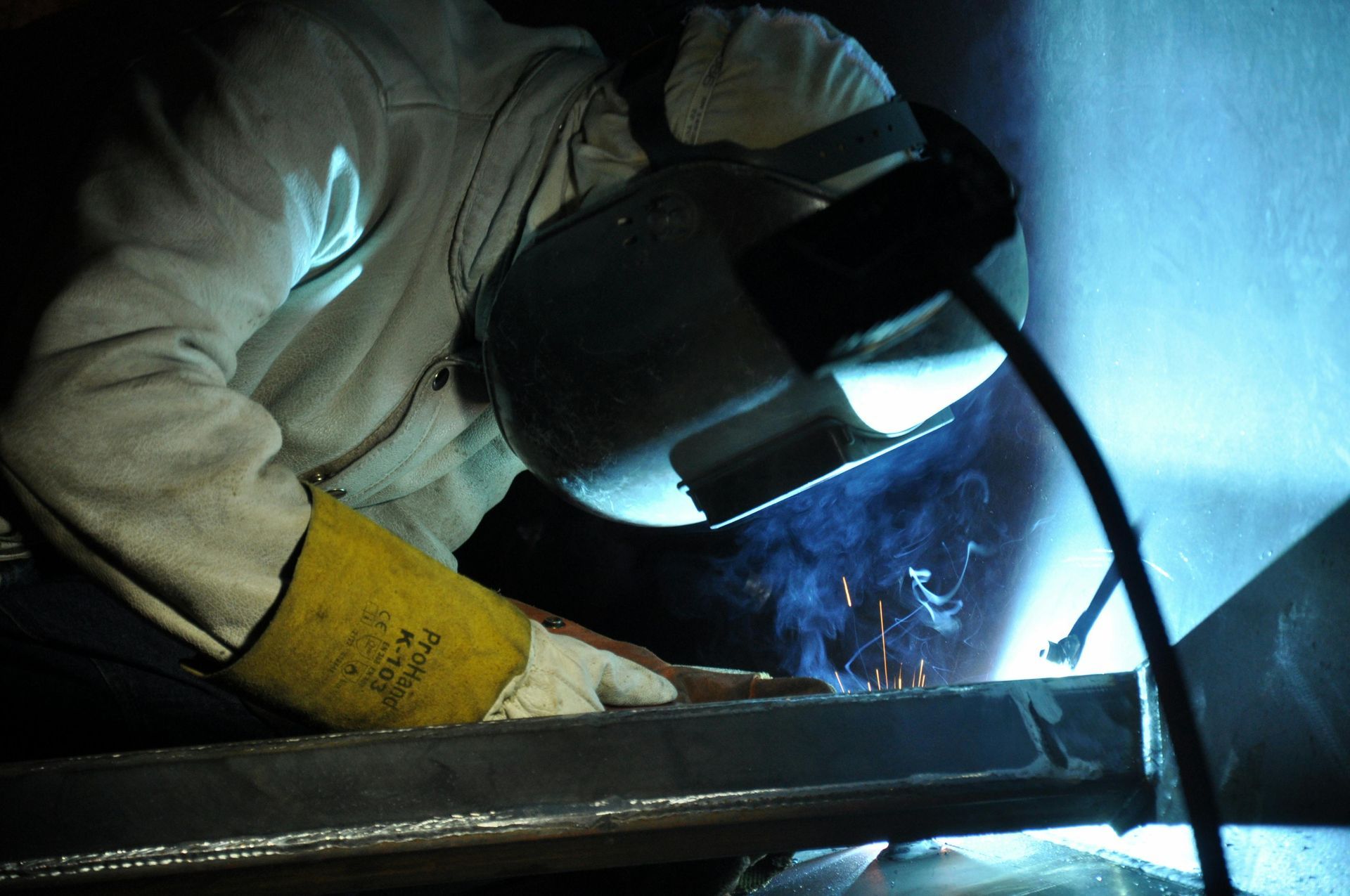 A welder in protective gear and a welding helmet works on a metal structure, emitting bright light and sparks.