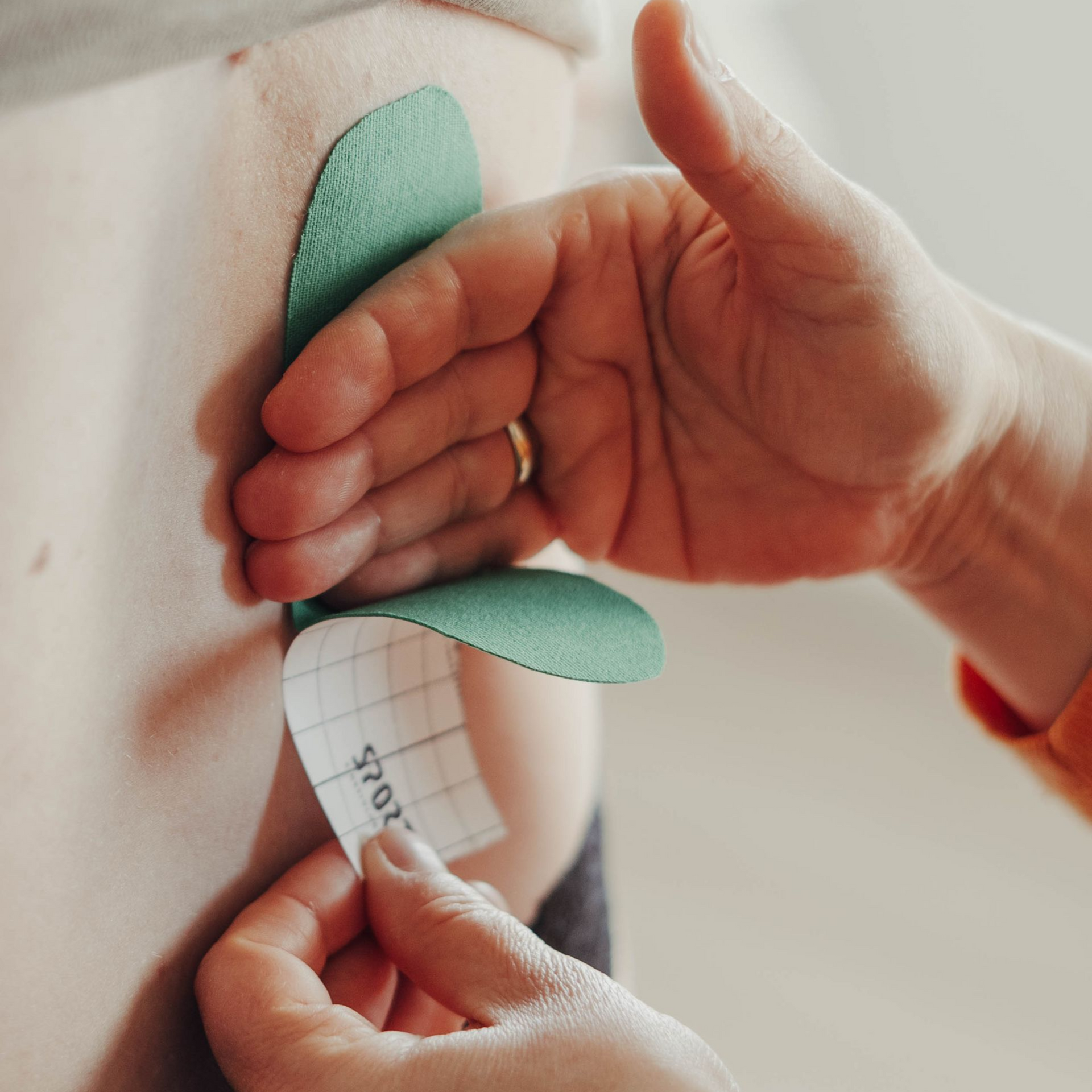 Der Rücken einer Frau wird mit grünem Kinesiologie-Tape umwickelt; eine Hand zieht die Schutzfolie ab.