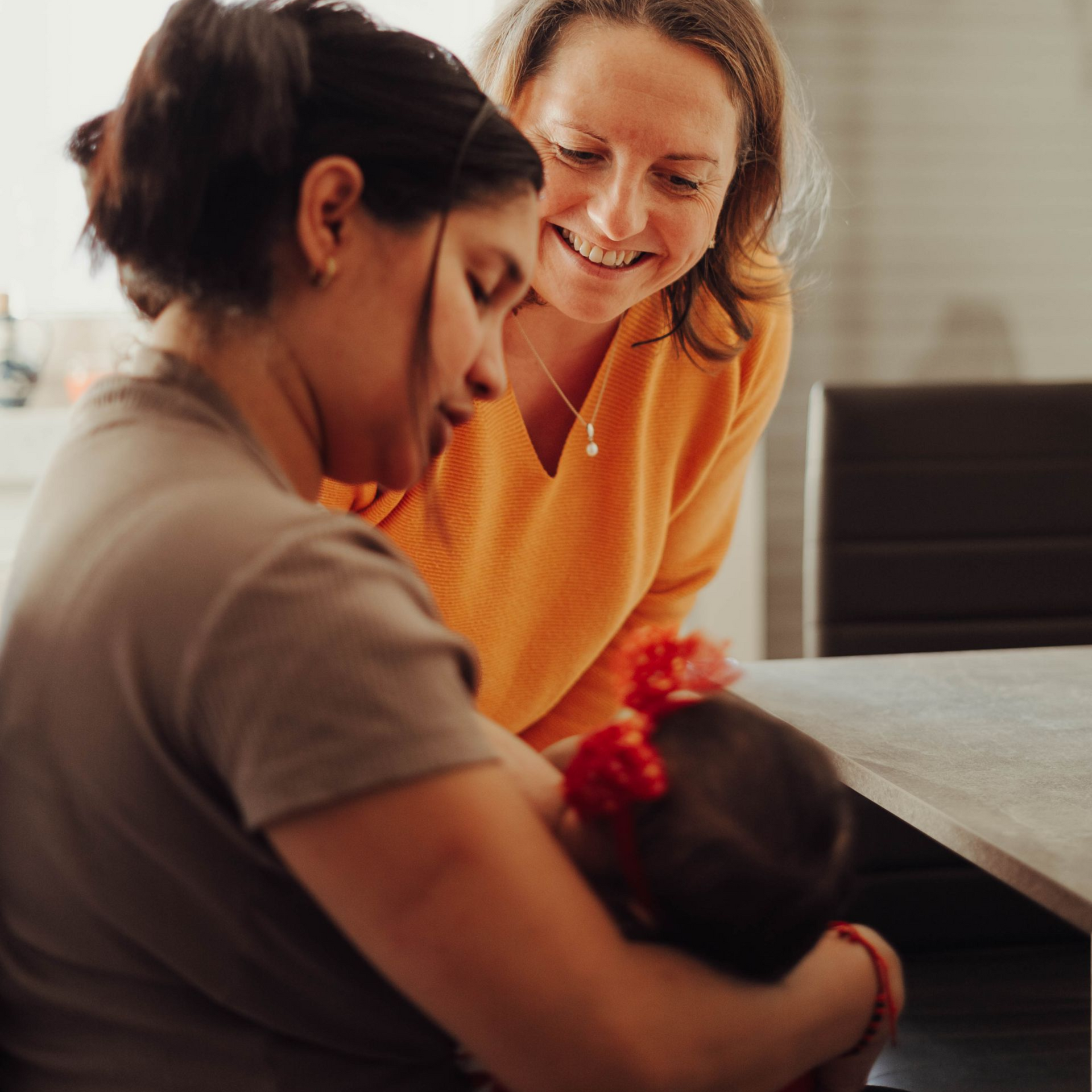 Eine Frau stillt ihr Baby; Stillberaterin Anja lächelt und schaut zu. Innenaufnahme, natürliches Licht.