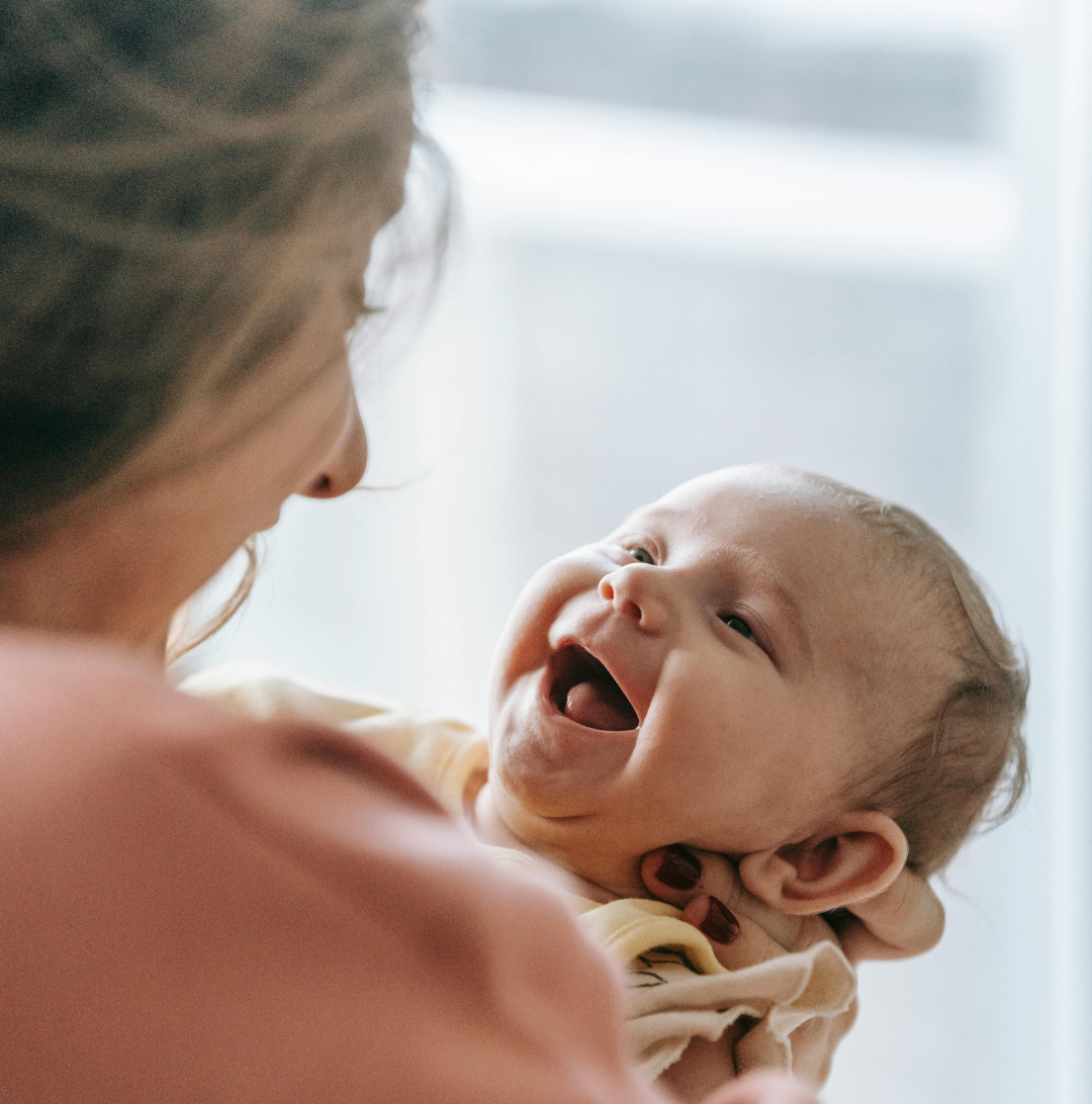 Frau hält ein lachendes Baby im Arm, drinnen. Das Baby trägt ein gelbes Outfit.