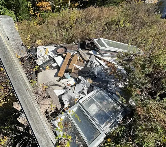 Pile of discarded debris and broken window frames in tall grass near a wooden post.