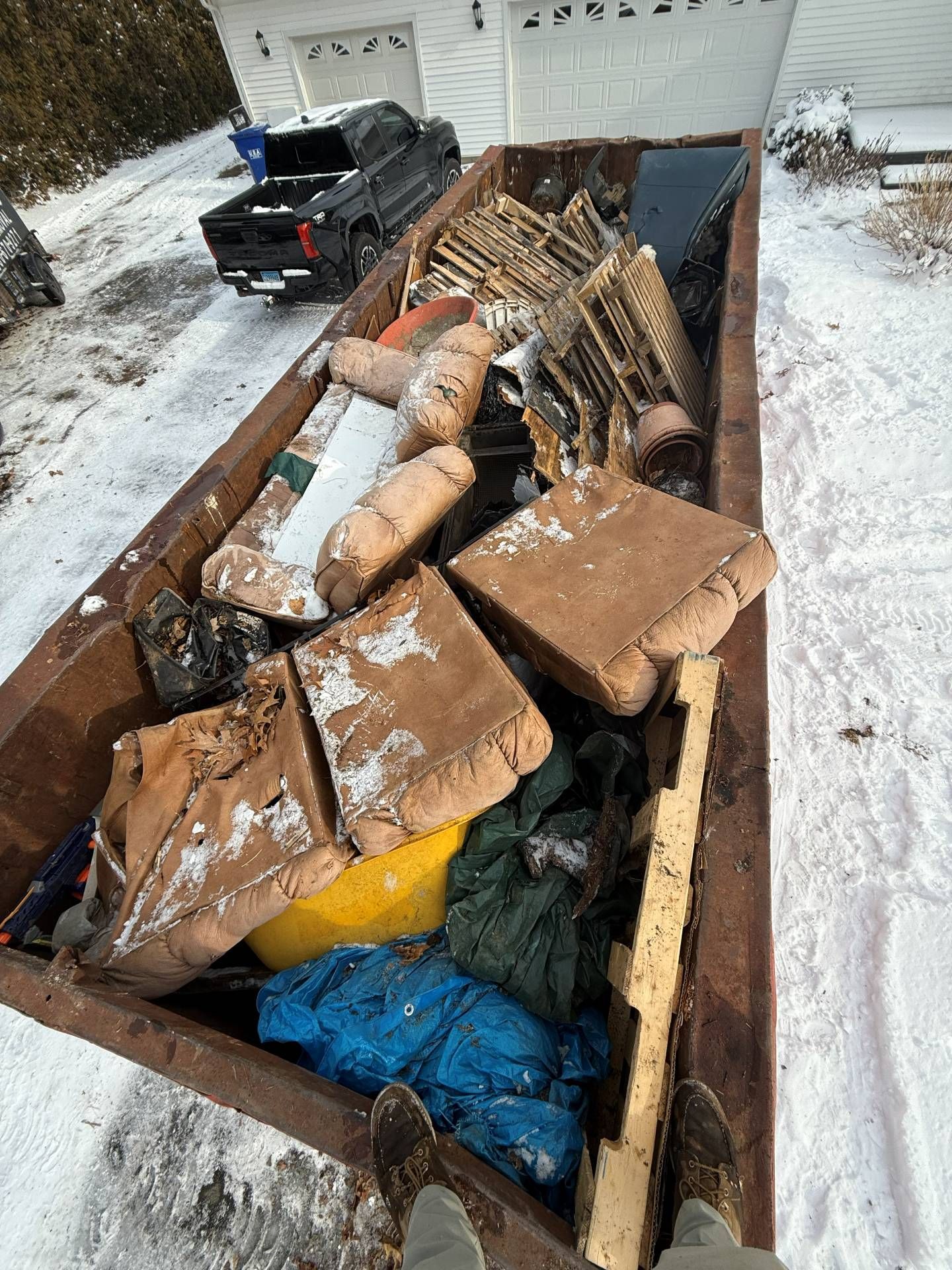 A dumpster overflowing with furniture on a snowy driveway, a black truck in the background.