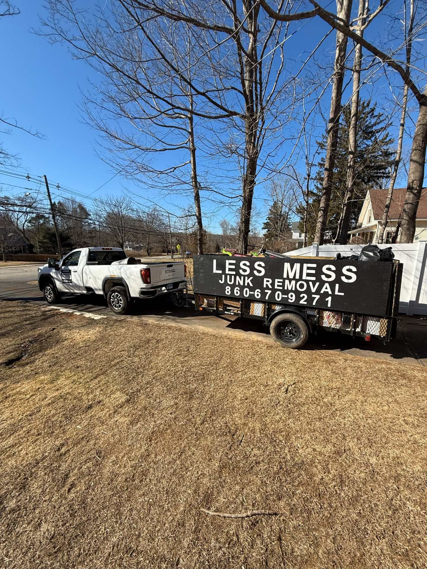 White truck towing a Less Mess Junk Removal trailer on brown grass, trees in the background, sunny day.