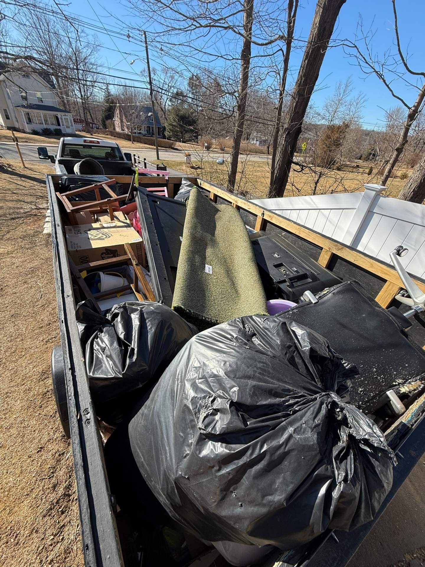 Trailer filled with debris: black trash bags, rug, wood, and other items in a sunny outdoor setting.