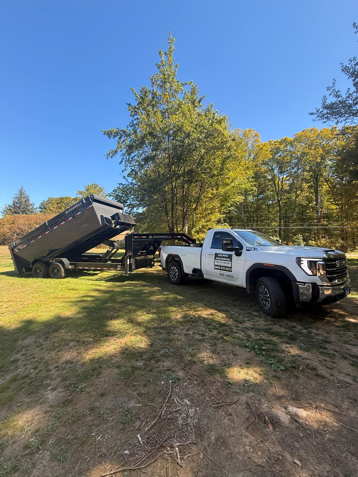 White pickup truck towing a black dump trailer on a grassy field with trees under a blue sky.