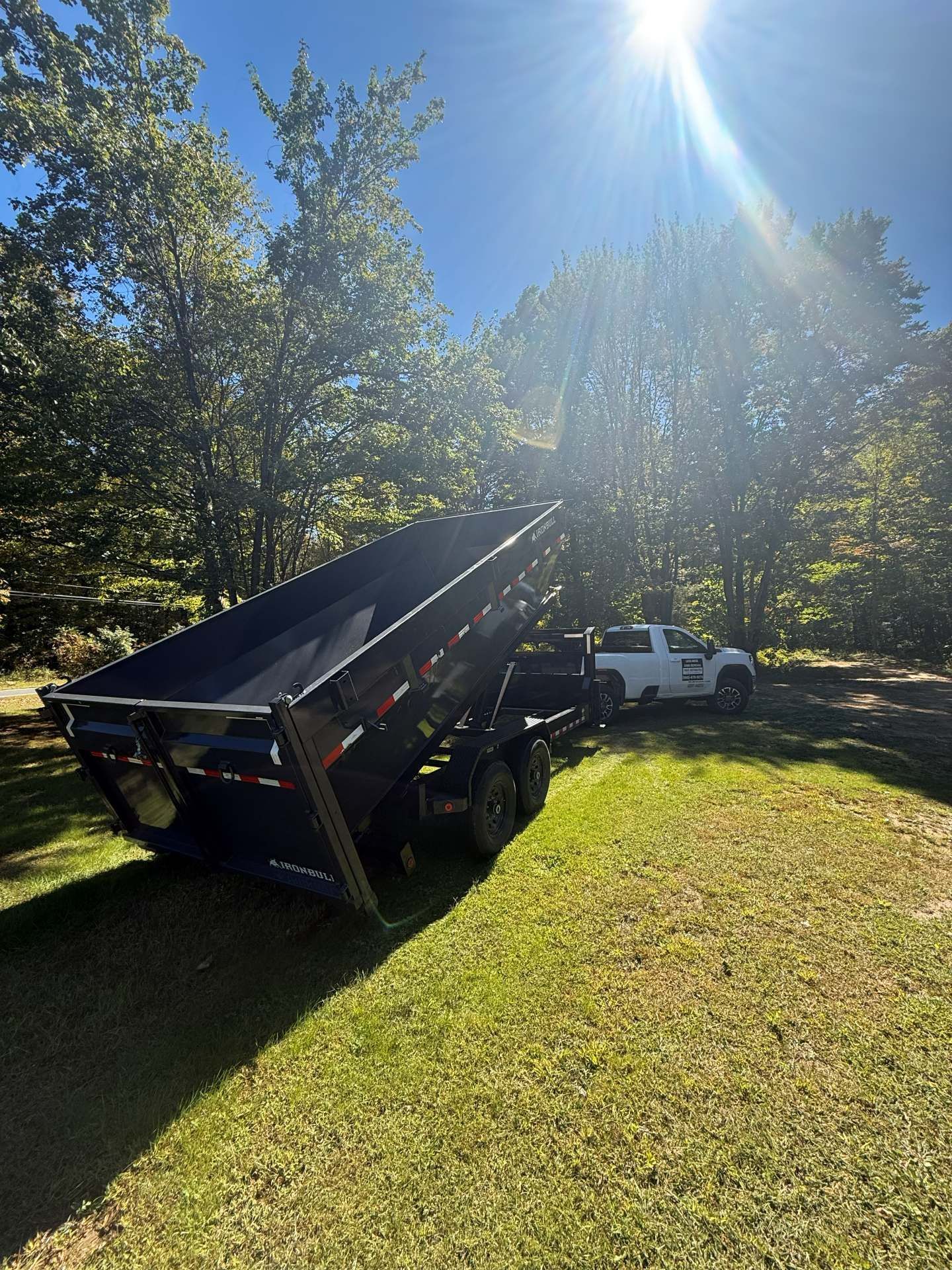 Dump trailer tilted up, being towed by a white truck on green grass with sun shining through trees.