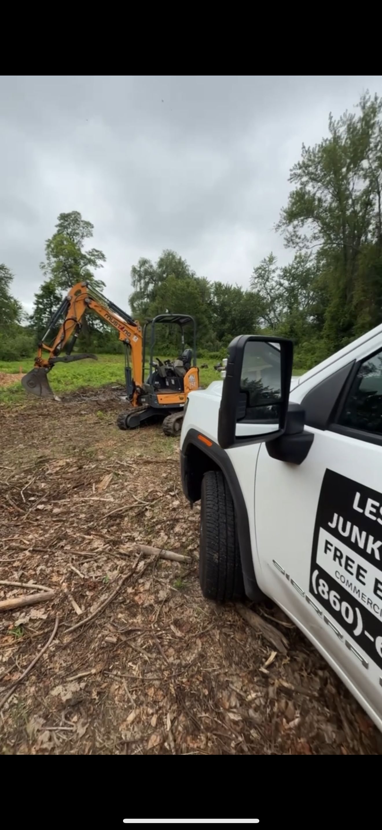 An excavator works in a field with a white truck in the foreground, cloudy sky overhead.