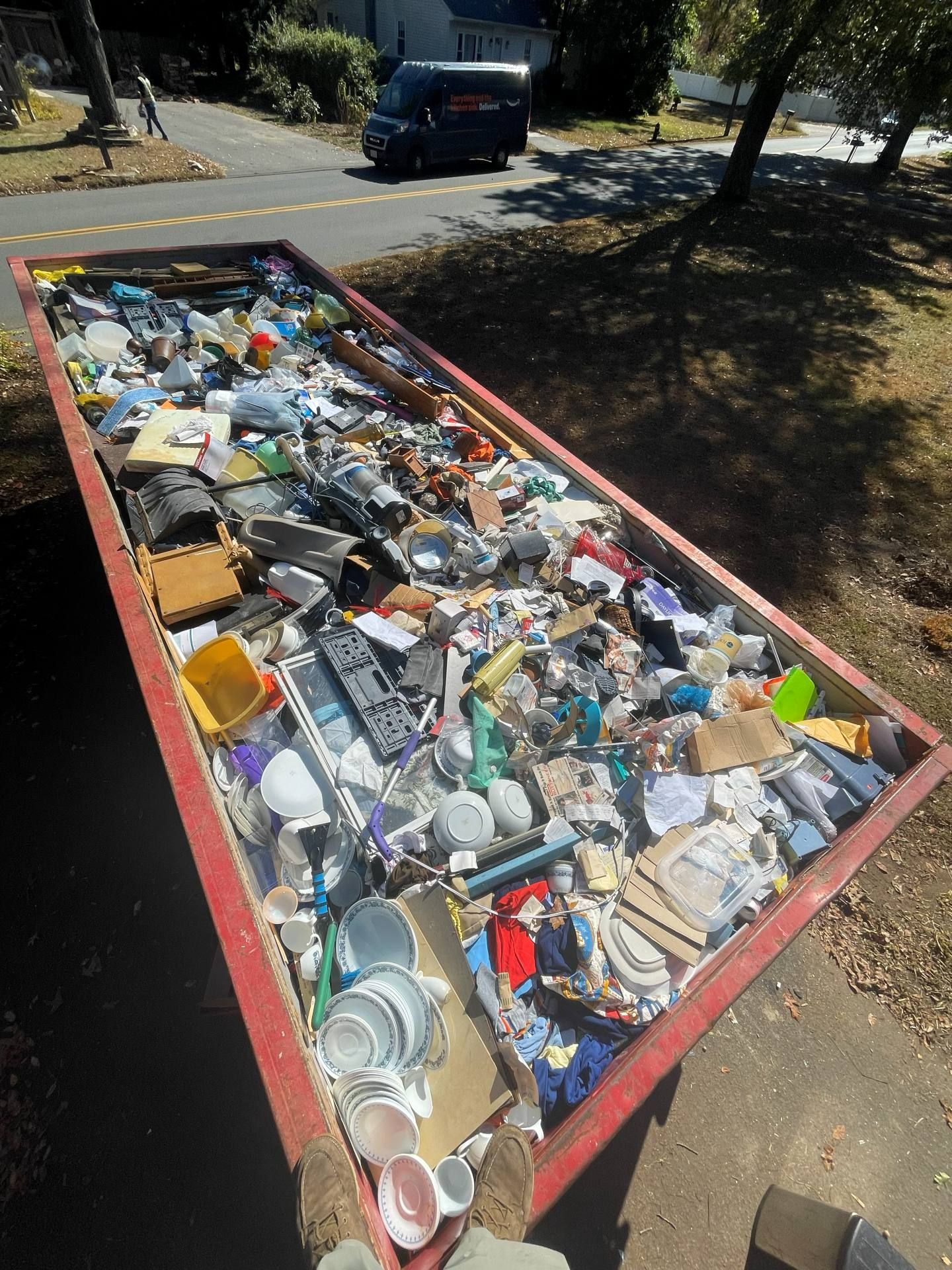 Red dumpster filled with trash on a sunny day; a van is visible in the background.