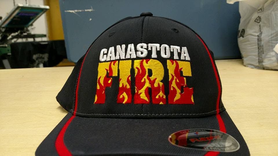 Black Canastota Fire cap with red-yellow flame text, resting on a table with a small metal badge on the brim