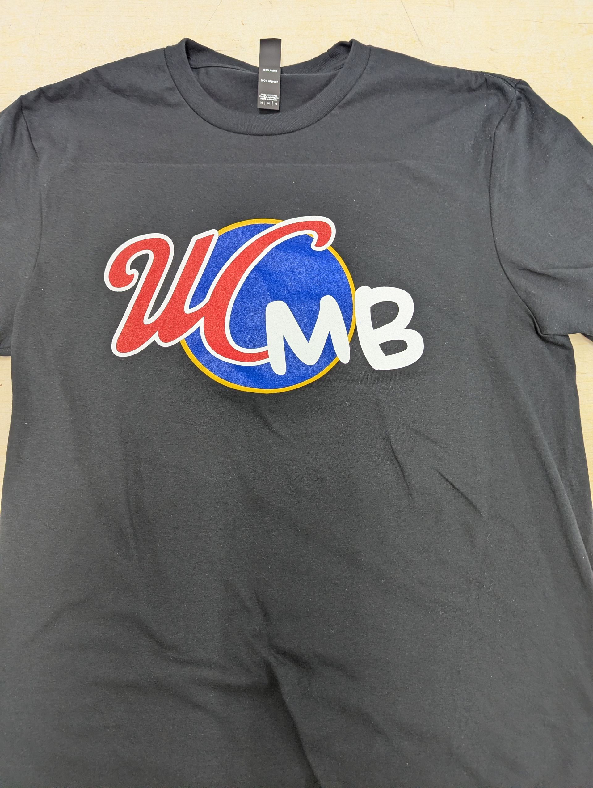 Dark gray T-shirt with a red, blue, and white UCMB logo centered on the chest