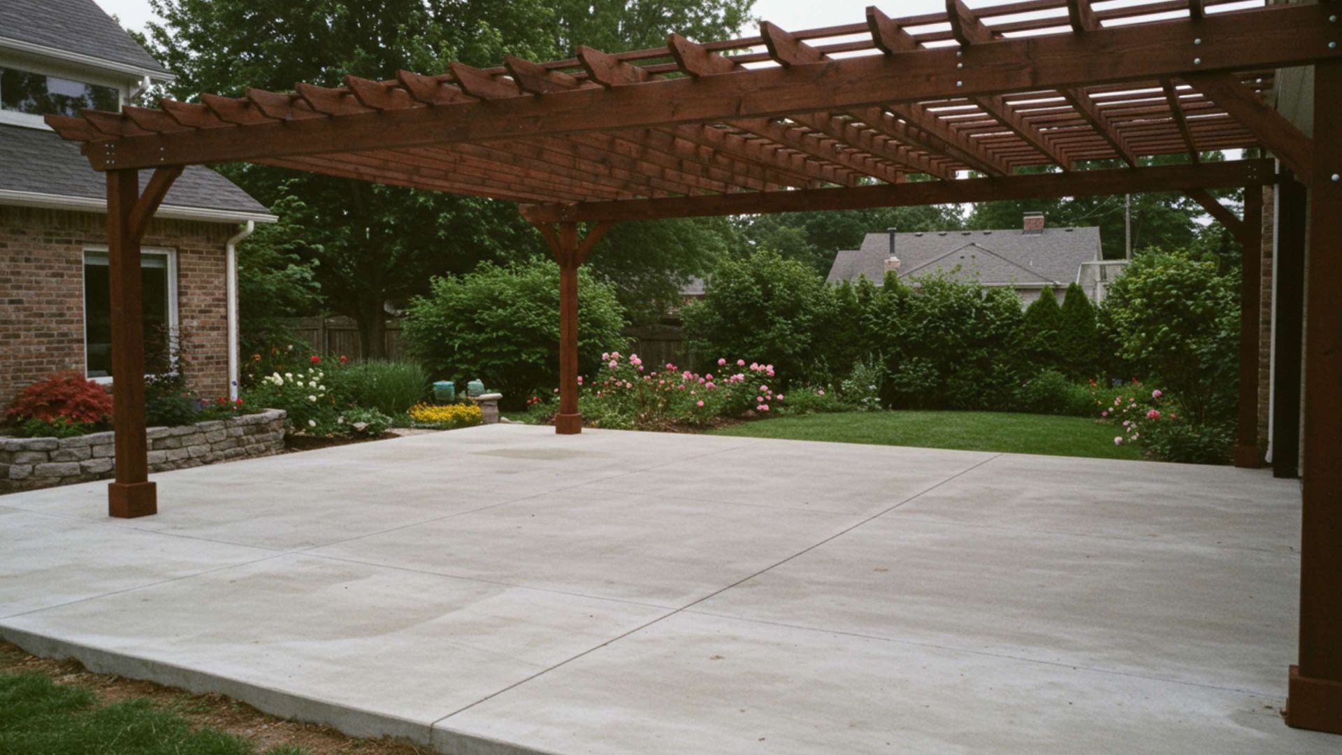 A wooden pergola stands over a large, light gray concrete patio next to a brick house and a lush green backyard.