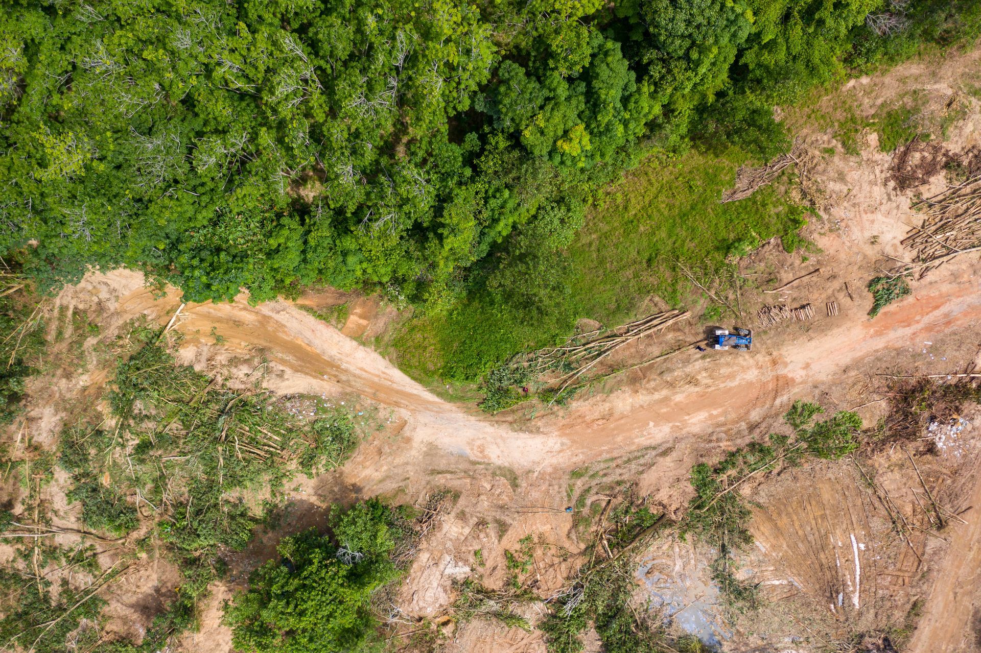 Aerial view of a dirt road curving through dense green forest, with a blue vehicle on the right side. Aerial view of a dirt road curving through dense green forest, with a blue vehicle on the right side.
