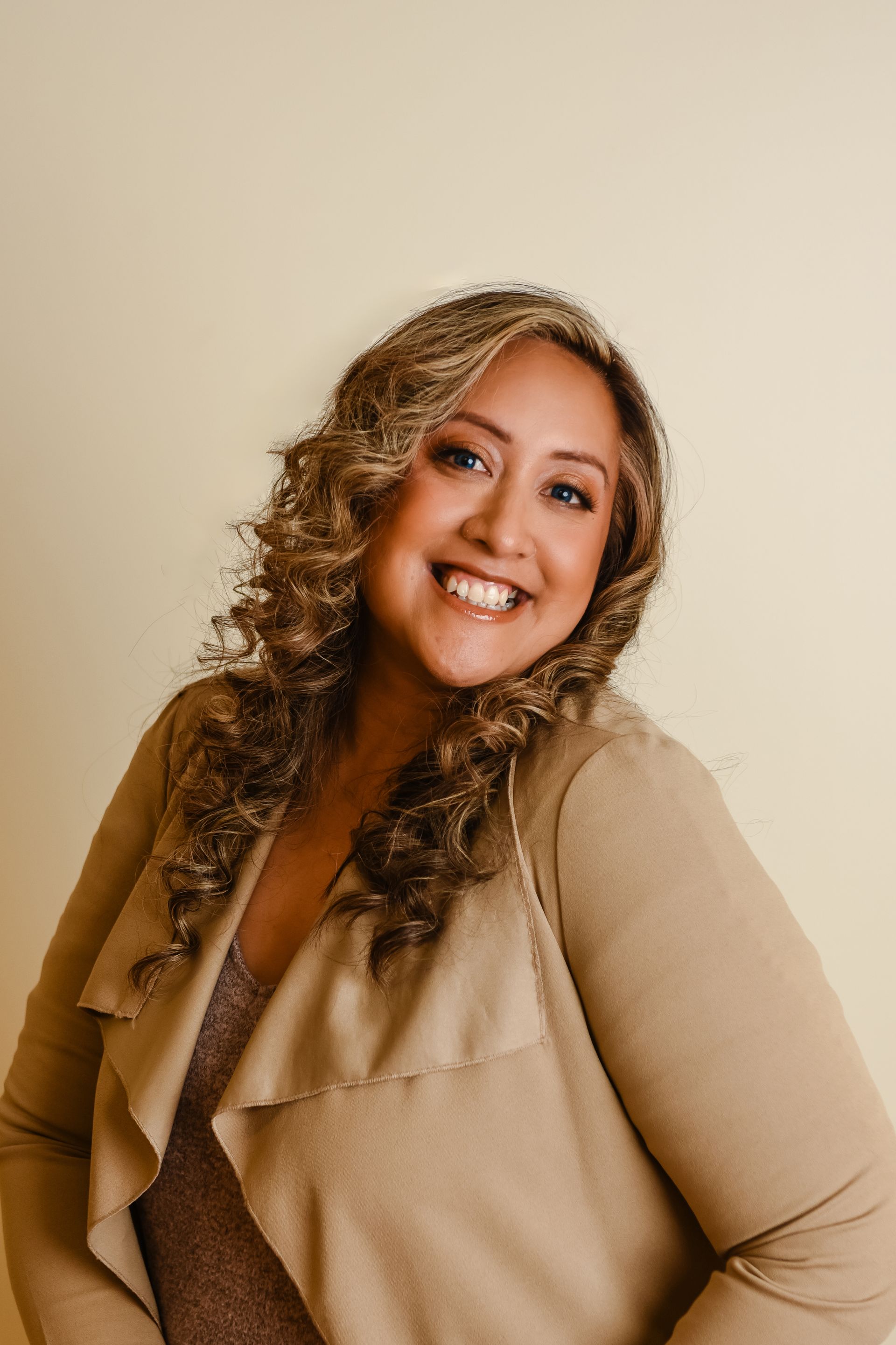 A smiling person with long, curly highlighted hair wearing a tan open-front cardigan against a light beige background.