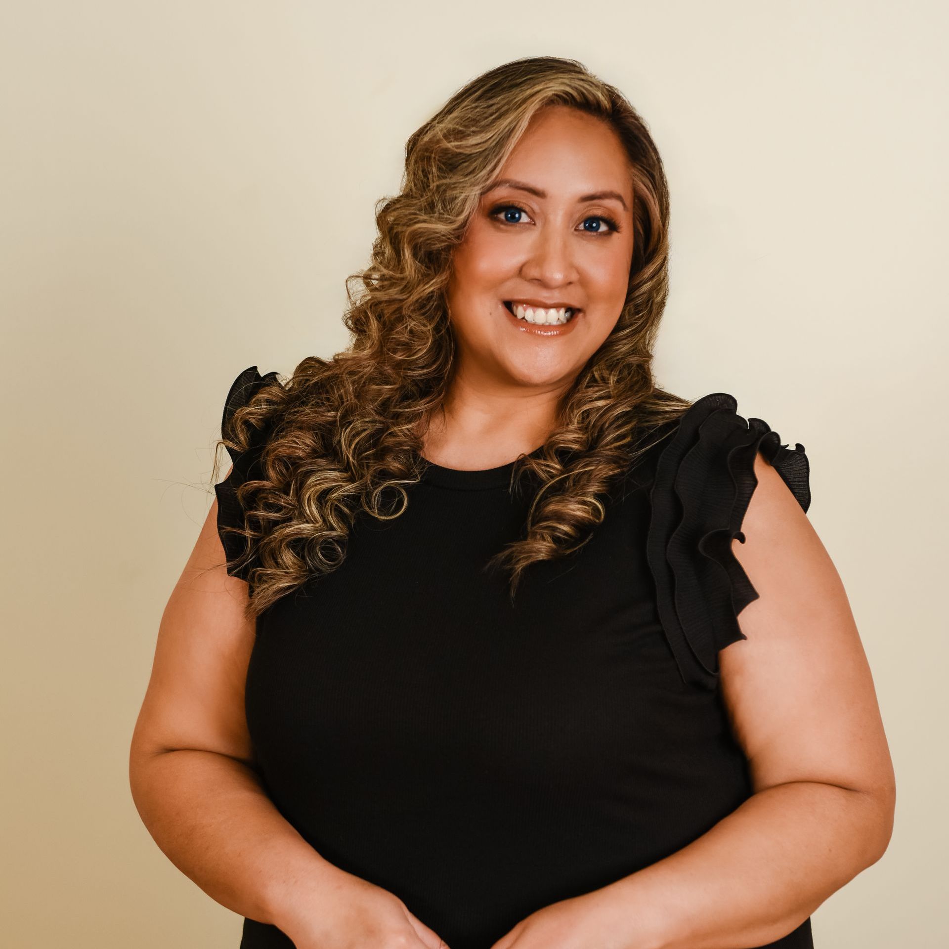 A smiling person with long, wavy brown hair wearing a black ruffled-sleeve shirt against a plain beige background.