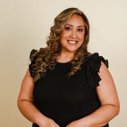 A smiling person with long, wavy brown hair wearing a black ruffled-sleeve shirt against a plain beige background.