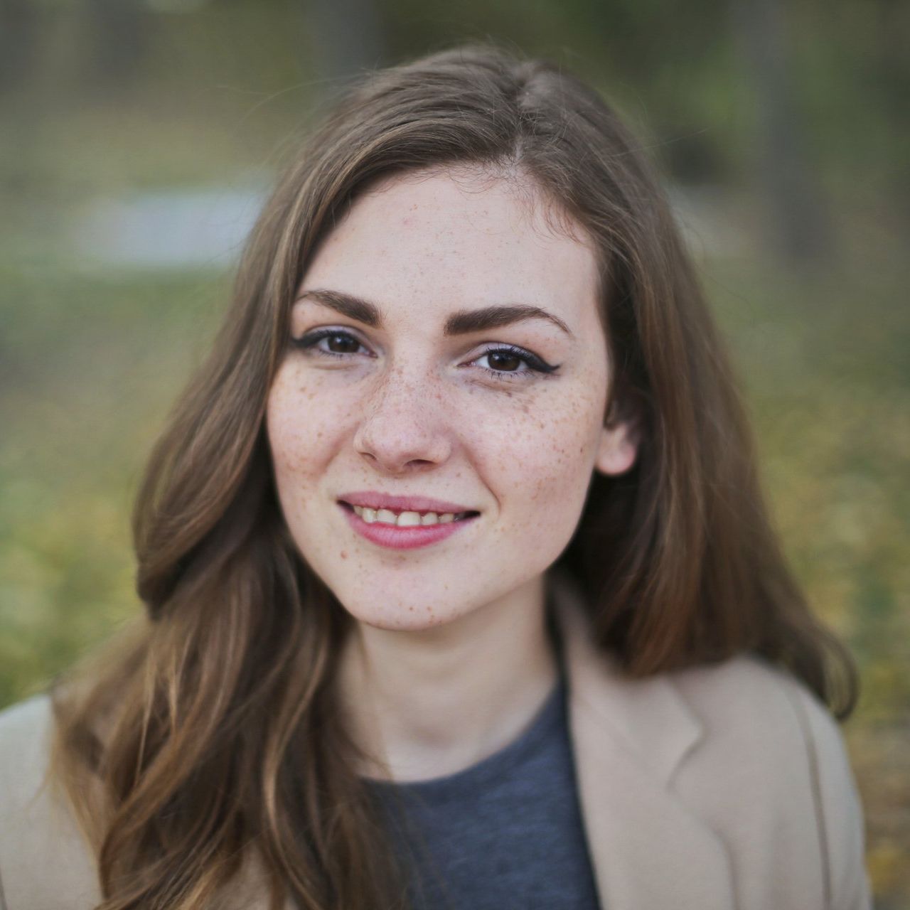 A person with long brown hair and freckles smiles softly, wearing a beige blazer, outdoors against a blurred background.