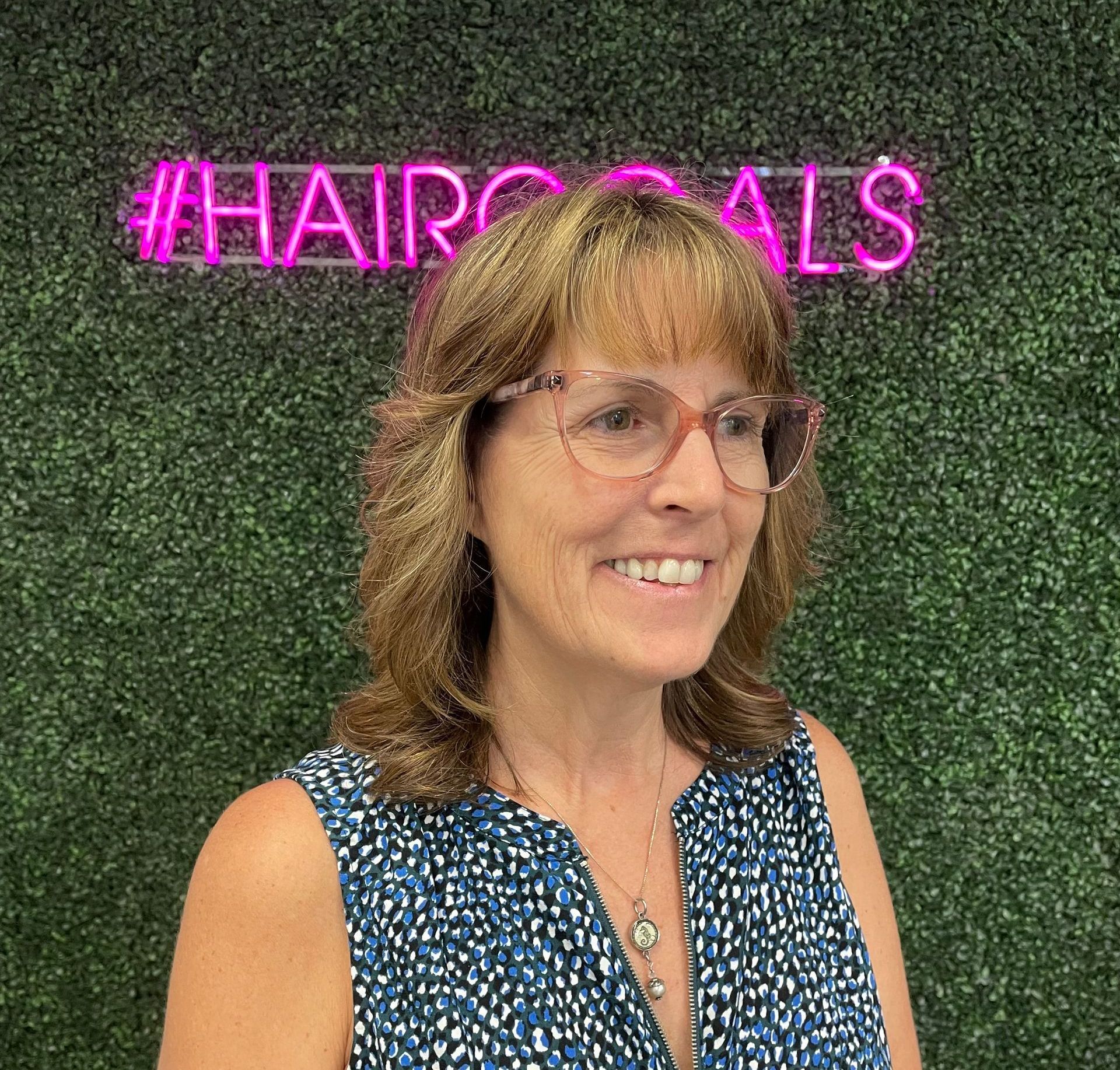 A woman wearing glasses and a blue shirt is smiling in front of a neon sign.