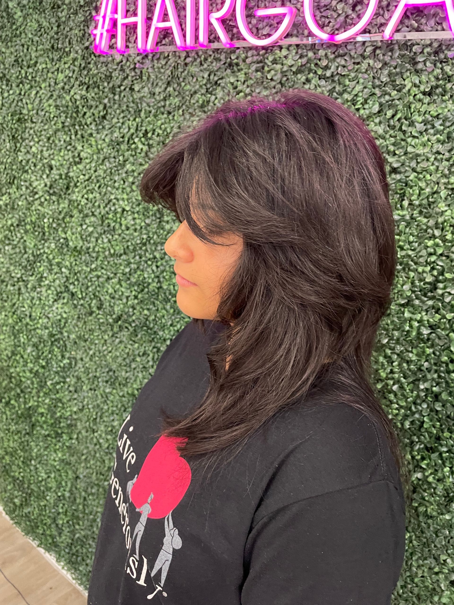 A woman with long hair is standing in front of a hedge wall.