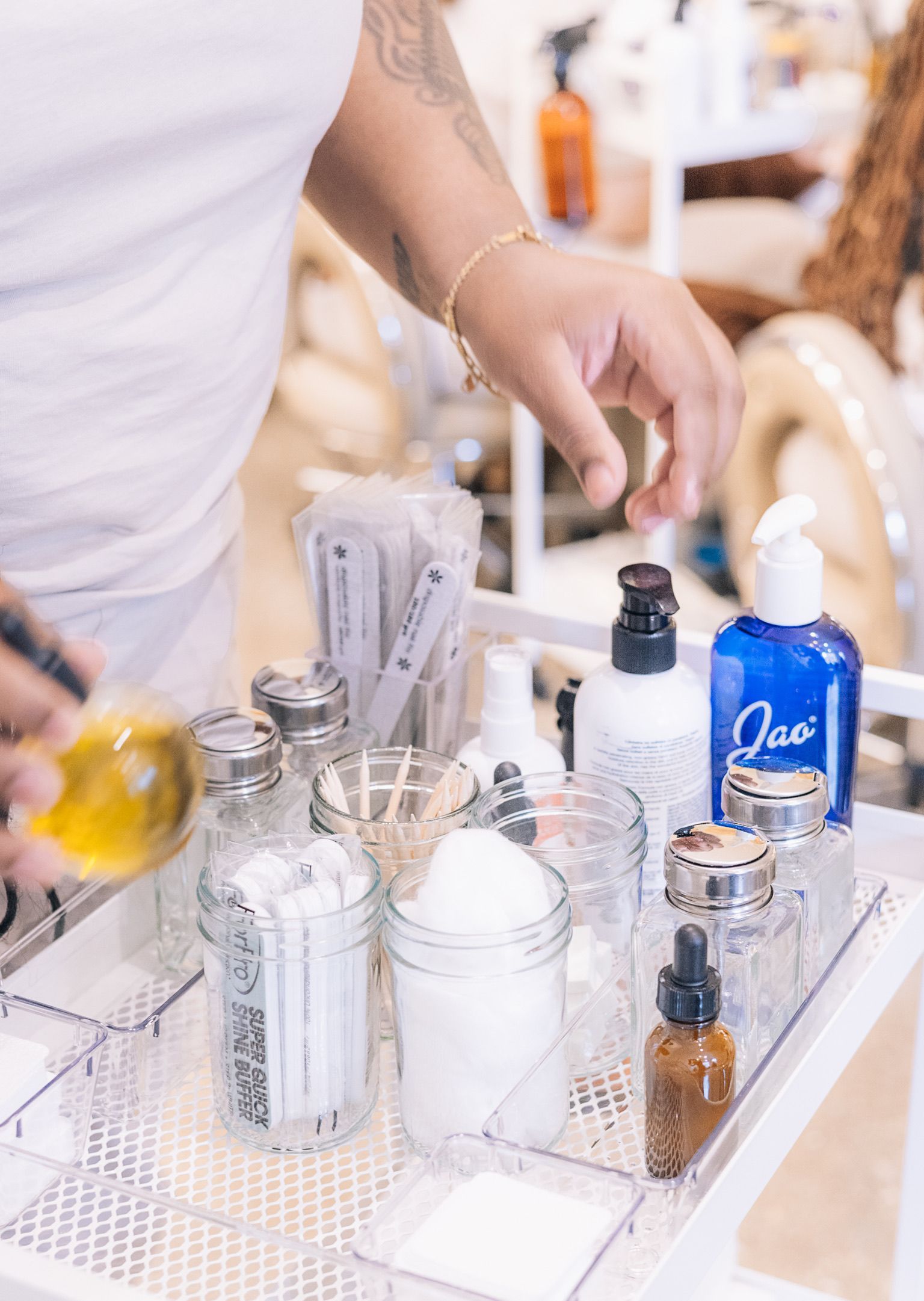 A person uses a small oil bottle over a salon cart filled with various bottles, jars of cotton, and nail care supplies.