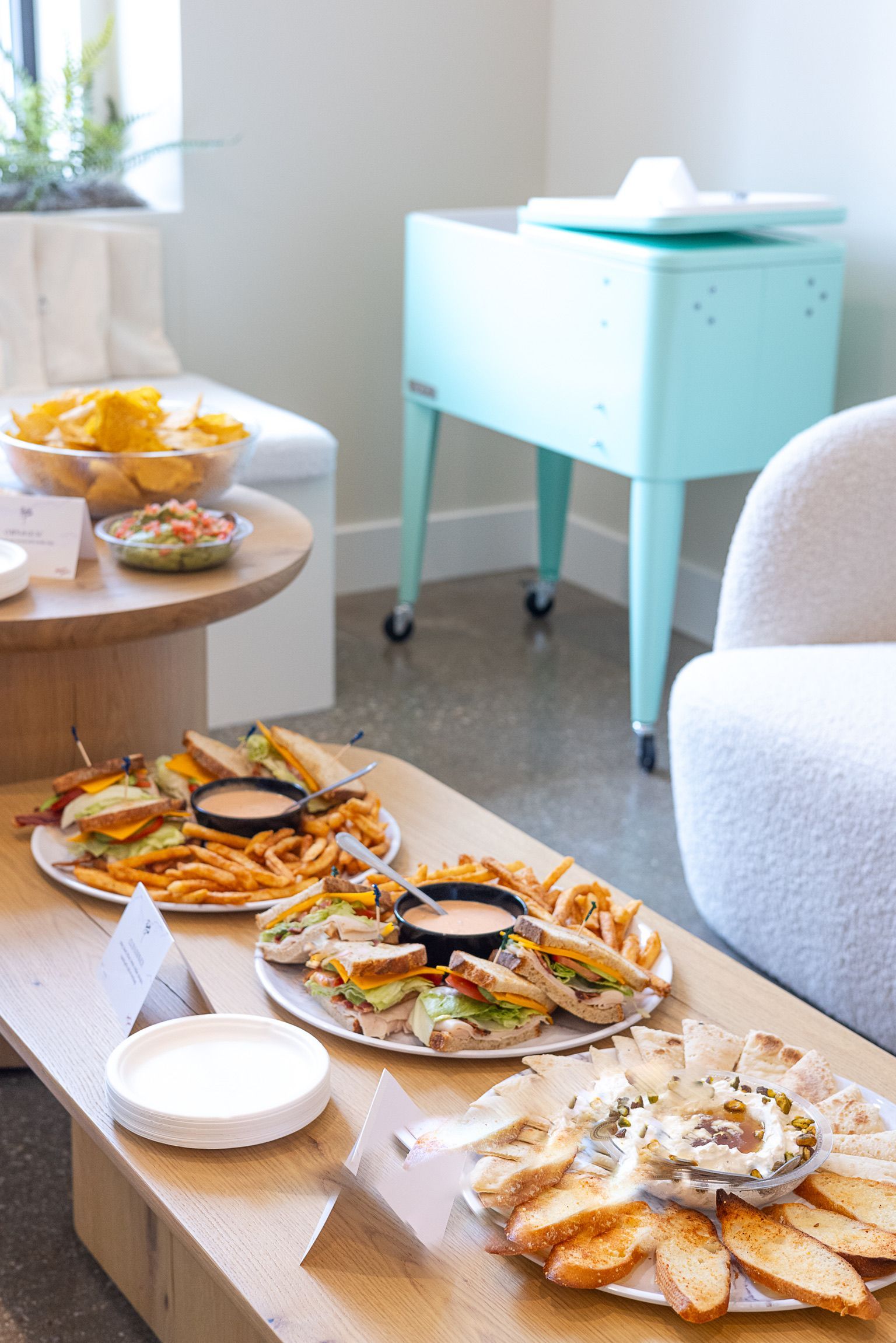Food platters including sandwiches, fries, and dip on a wooden table, with a light blue vintage cooler in the background.