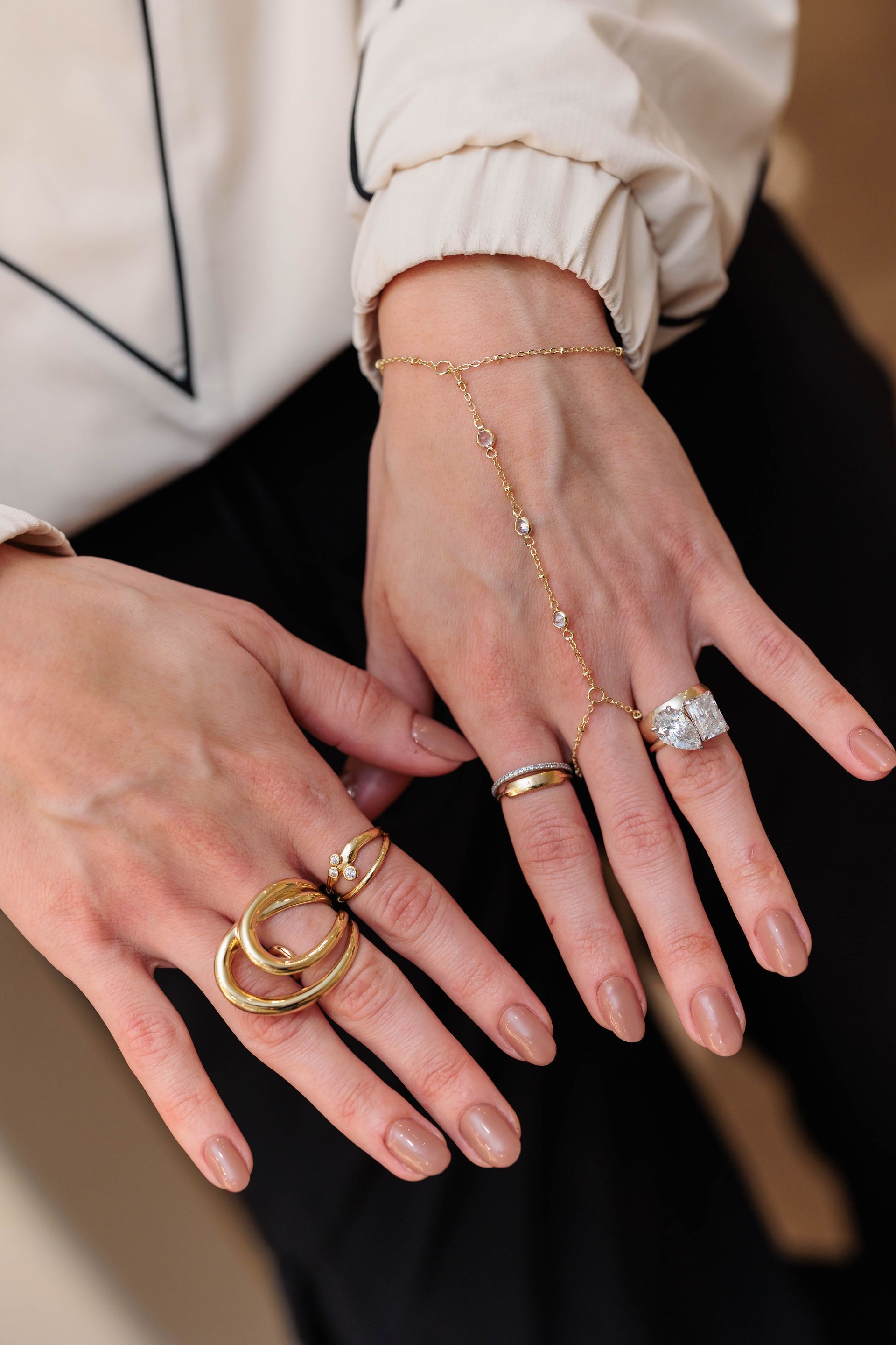 Hands wearing multiple gold rings and a delicate hand chain connecting a bracelet to a finger ring.
