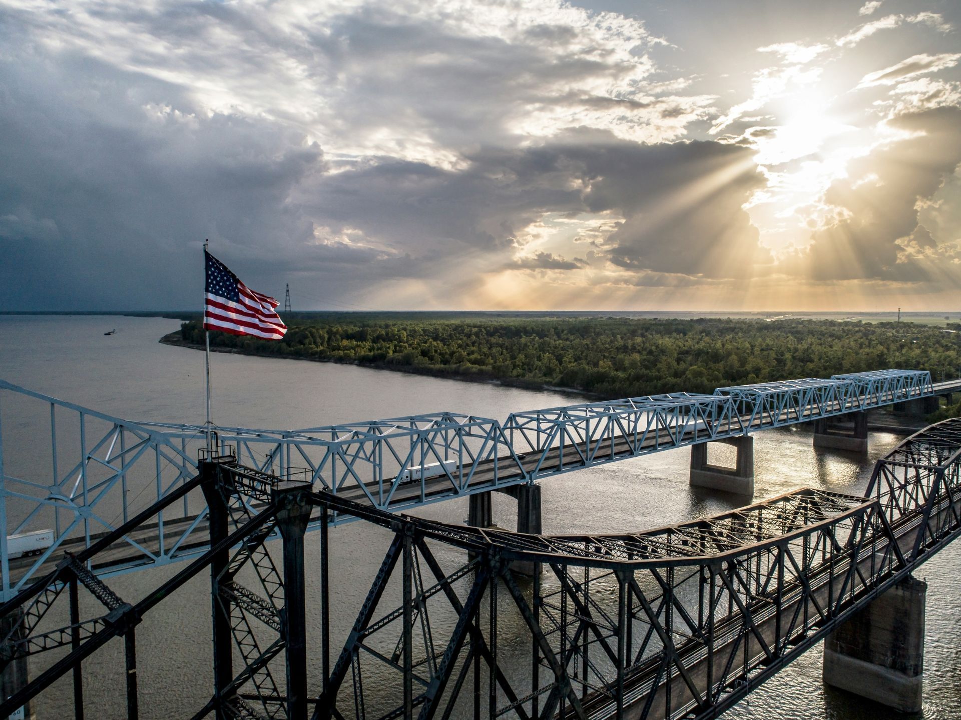 American flag on a bridge over water at sunset, with dramatic clouds and sunlight breaking through.