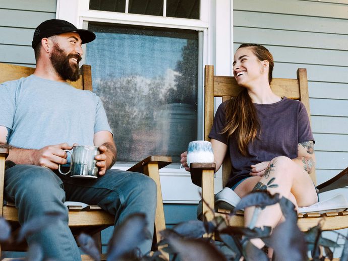 Two people sitting on a porch, smiling and talking over coffee with a dog in the foreground