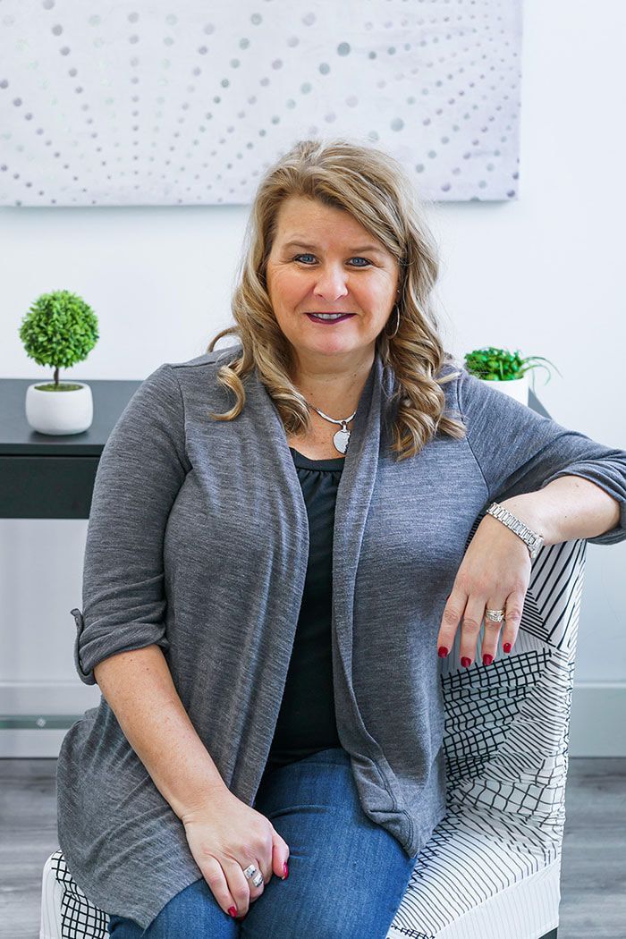 Woman seated on a patterned chair in a bright office, smiling beside small potted plants.