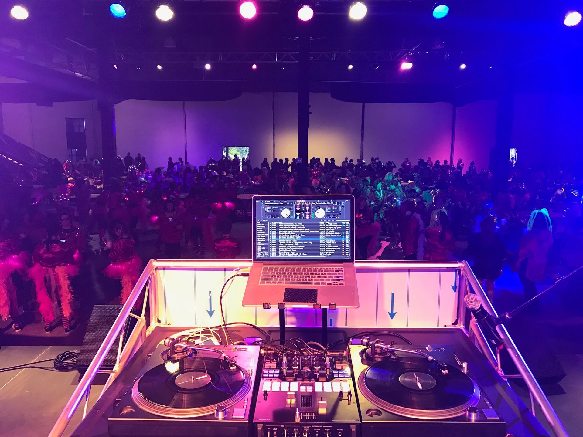 DJ booth facing a packed, purple-lit concert crowd with turntables and a laptop screen in view