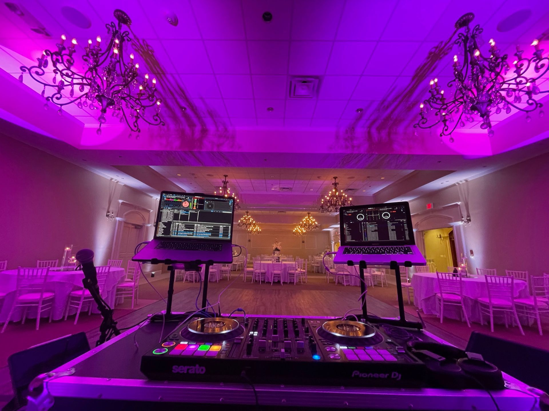 Purple-lit ballroom with DJ booth, twin monitors, chandeliers, and rows of white chairs at the sides