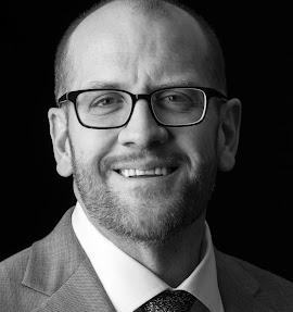 Smiling bald man wearing glasses and a suit against a dark background