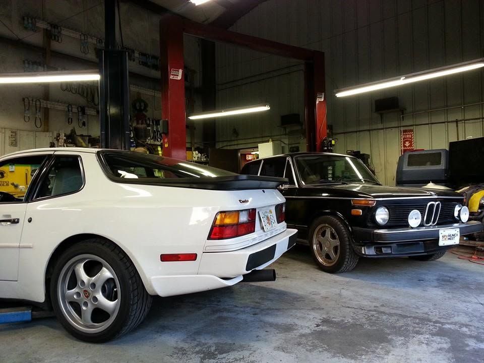 Two classic cars in a garage: a white Porsche 911 and a black BMW with rally lights. | Tony's European Auto Service