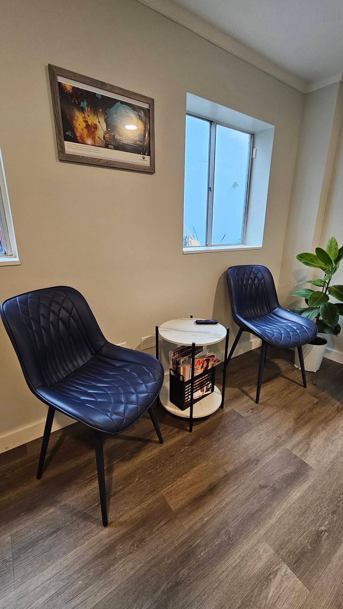 Small waiting area with two black chairs, a round side table, wall art, a window, and a plant. | Tony's European Auto Service