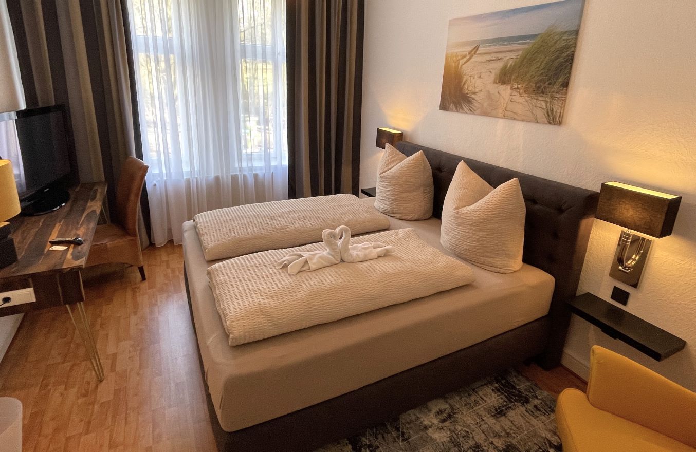 Hotelzimmer mit Bett, Schreibtisch und Fenster. Beige Bettwäsche, braune Vorhänge und Kunstwerke.