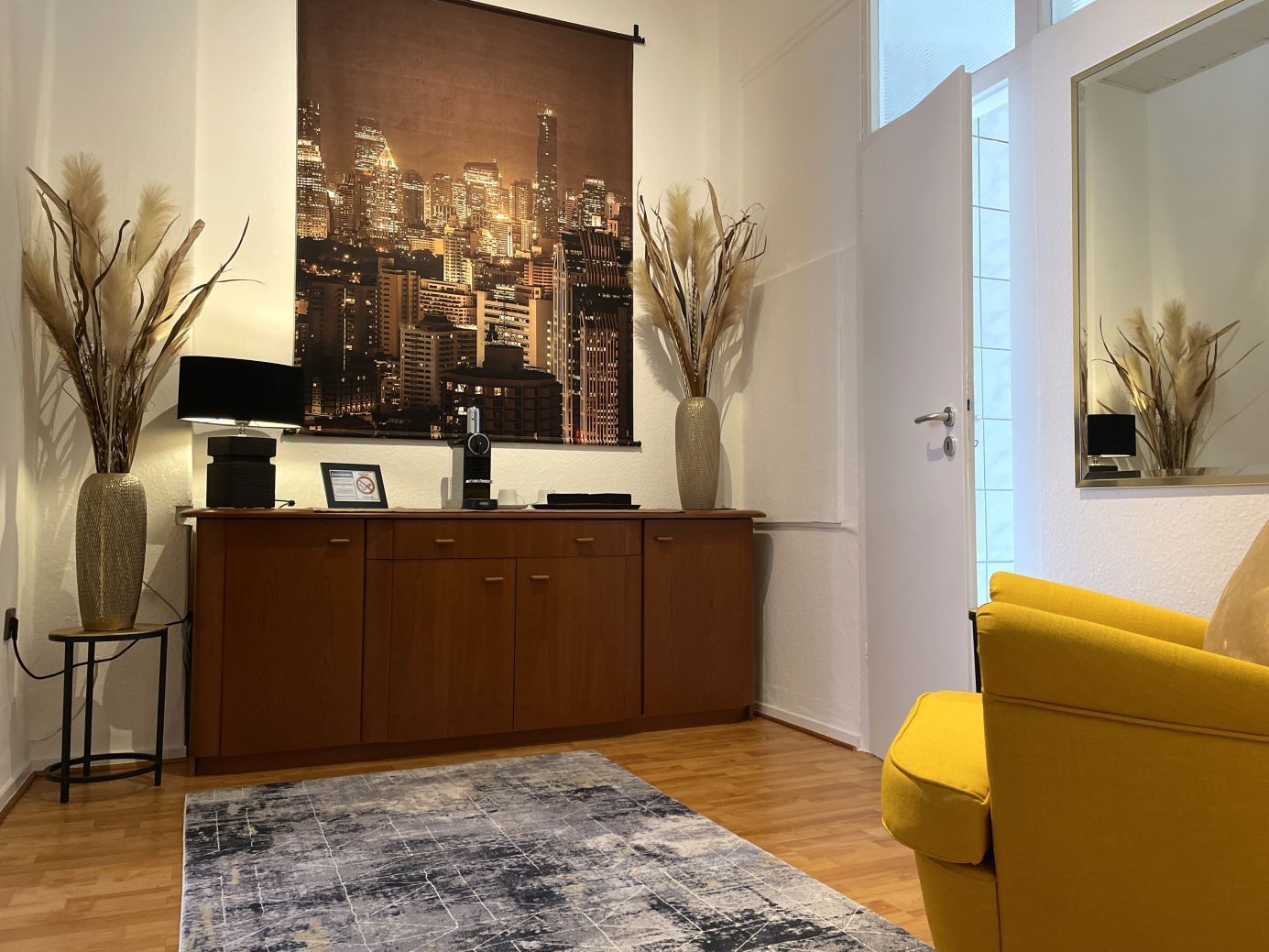 Ein Zimmer mit einem Schrank, einem Bild einer Stadt bei Nacht, hohen Vasen mit Zierpflanzen und einem gelben Stuhl.