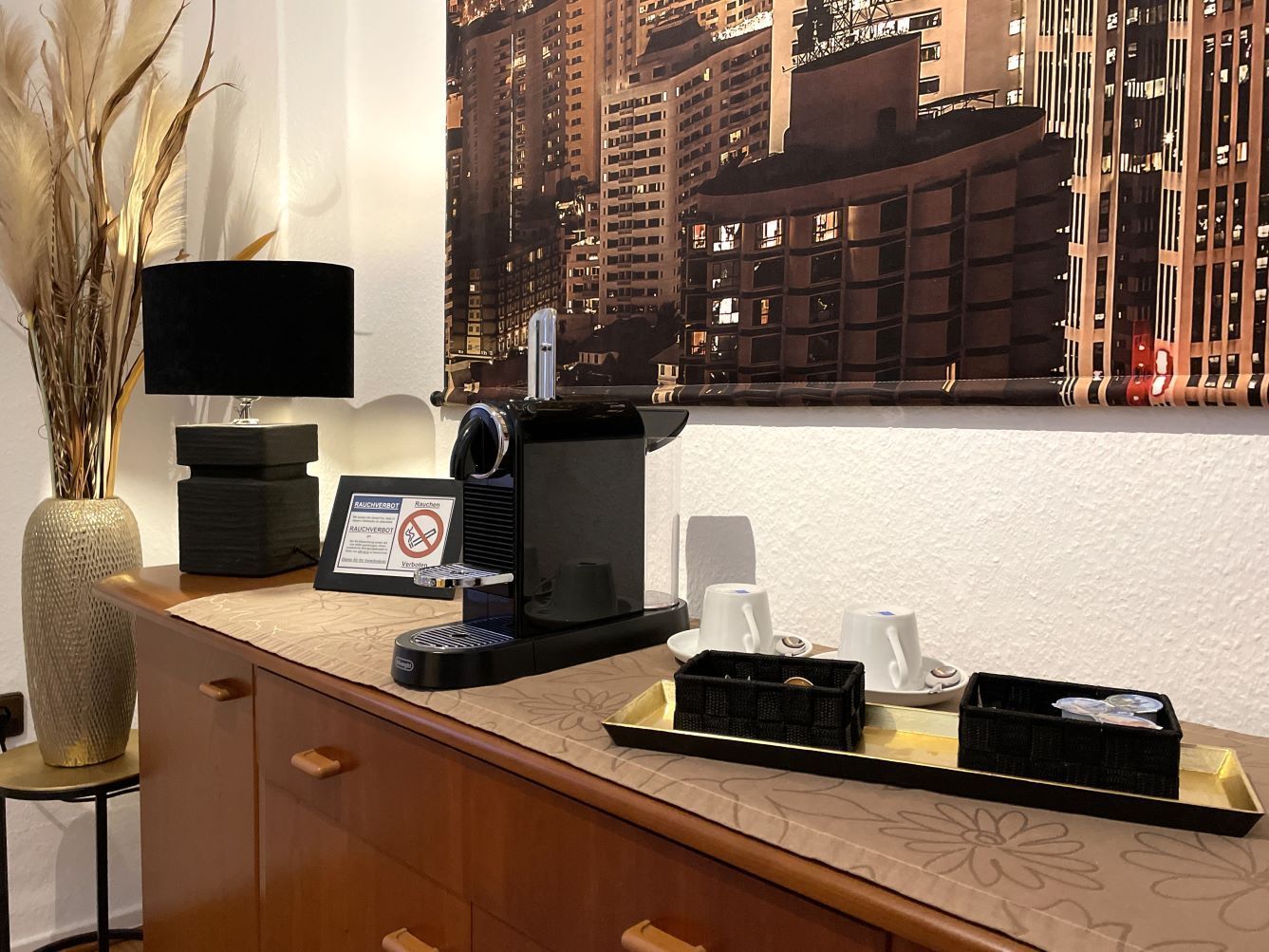 Kaffeestation auf einem Holzschrank, mit Lampe, Kaffeemaschine, Tassen und einem Stadtpanorama-Bild.