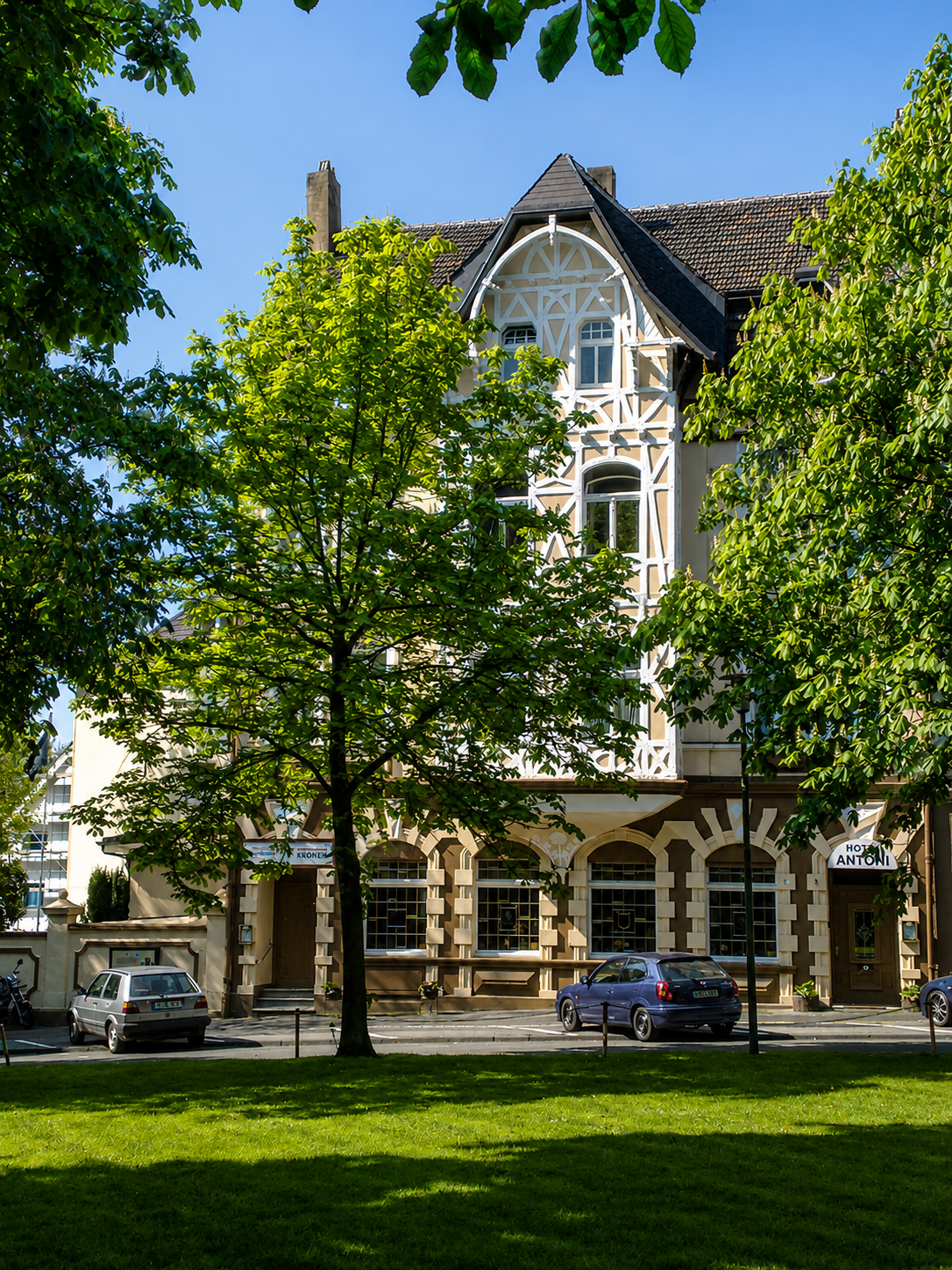 Zweistöckiges Gebäude mit reich verzierter Fassade, davor Bäume und geparkte Autos, klarer Tag.