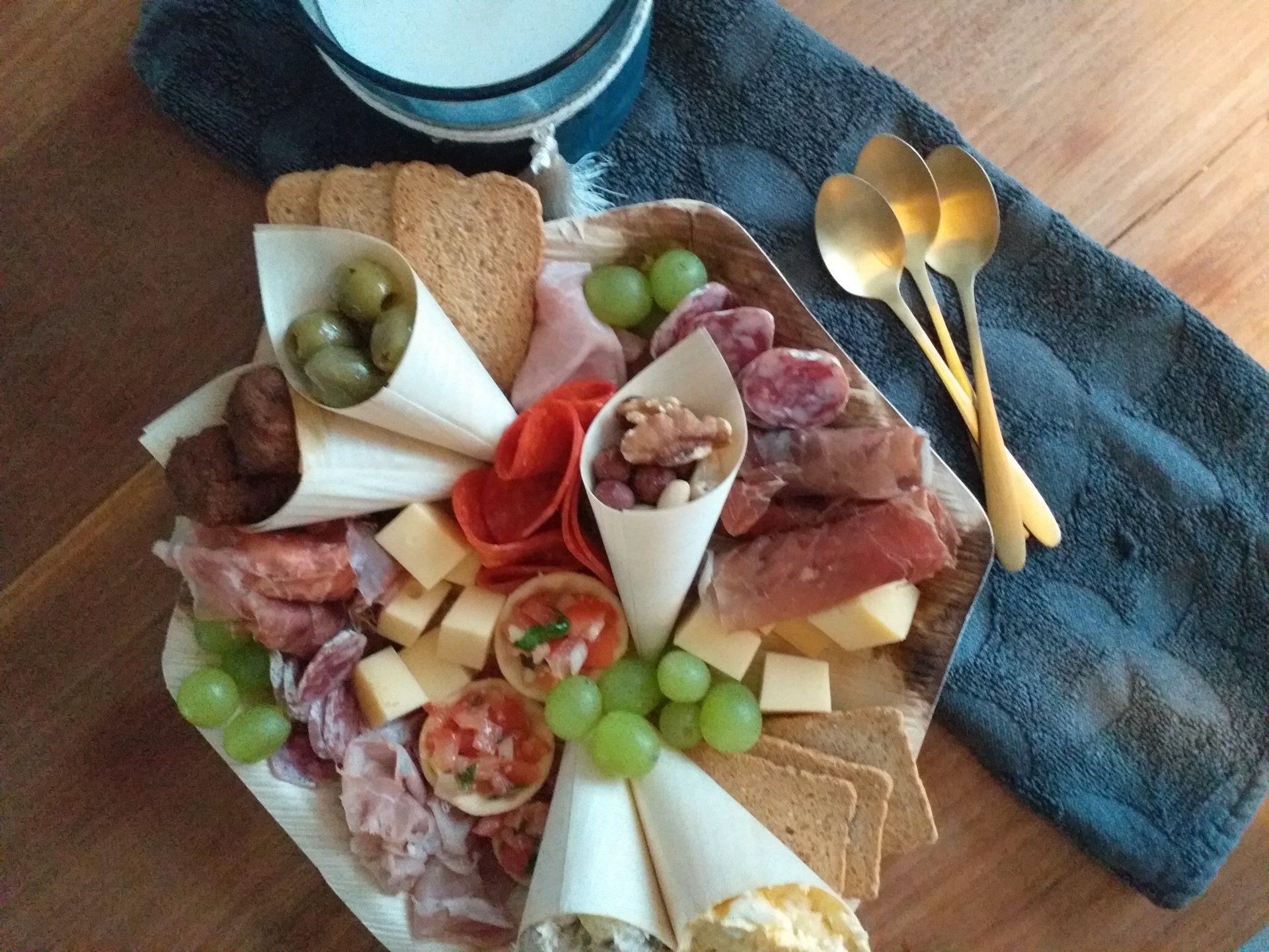 Een charcuterieplank met vleeswaren, kazen, crackers, olijven en noten in papieren puntzakken op een donkerblauw tafelkleed, naast gouden lepels.