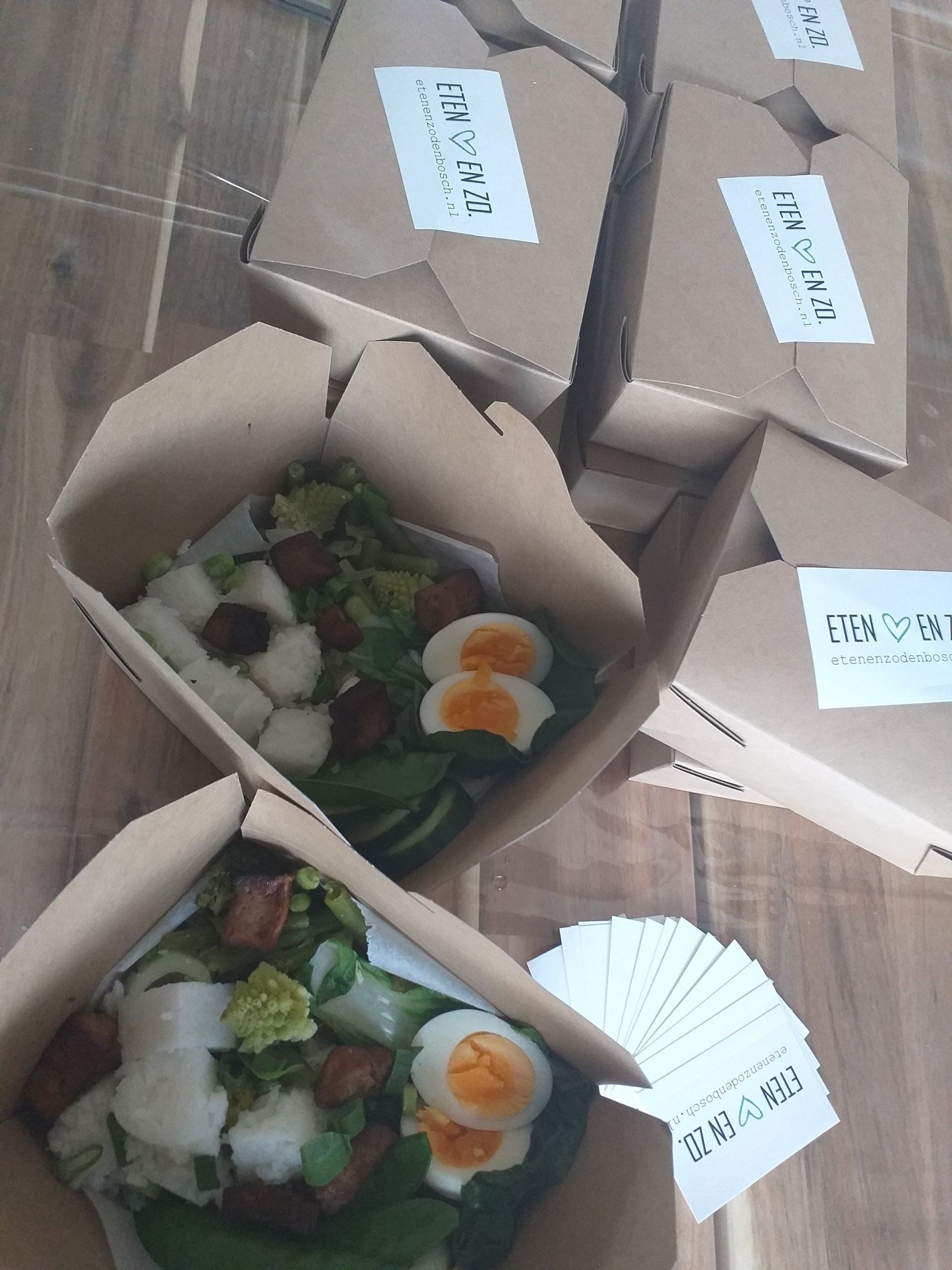 Op een houten ondergrond staan ​​verschillende kartonnen afhaalbakjes gevuld met salade, hardgekookte eieren en geroosterde blokjes.