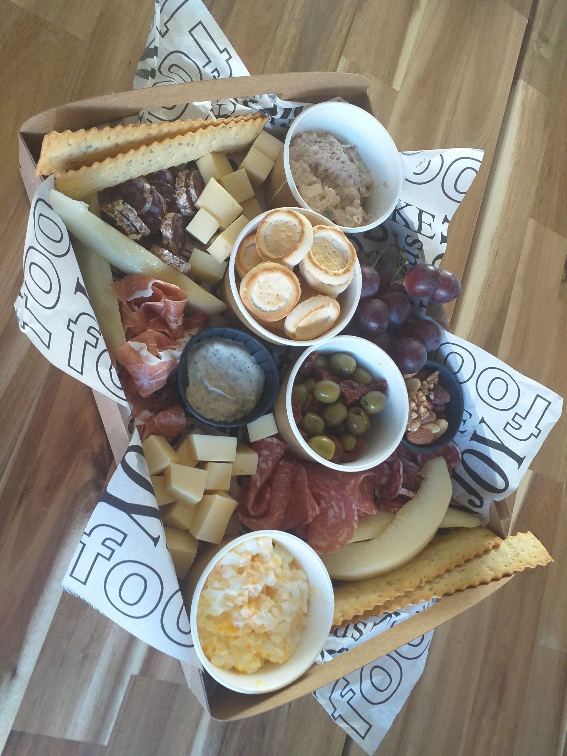 Een charcuteriebox op een houten ondergrond, gevuld met kazen, vleeswaren, crackers, druiven, olijven en kleine bakjes dipsaus.