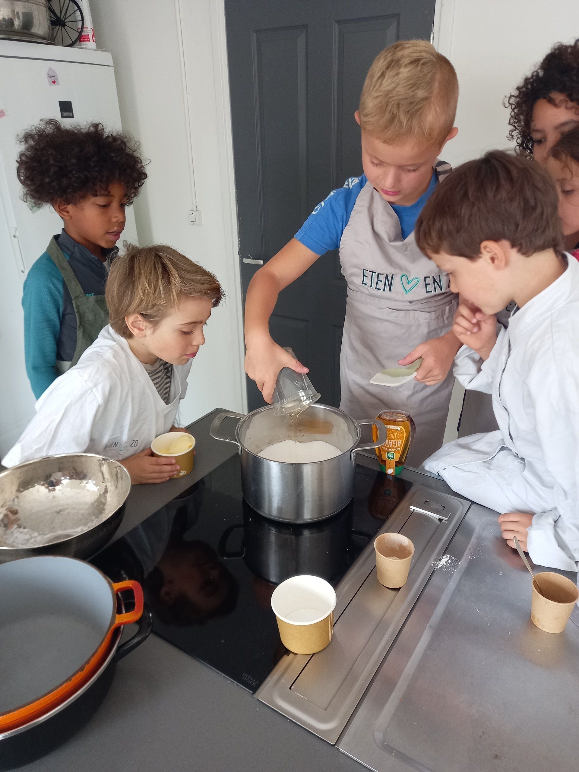 Kinderen met schorten om koken samen bij een fornuis en gieten ingrediënten in een pan, terwijl anderen toekijken.