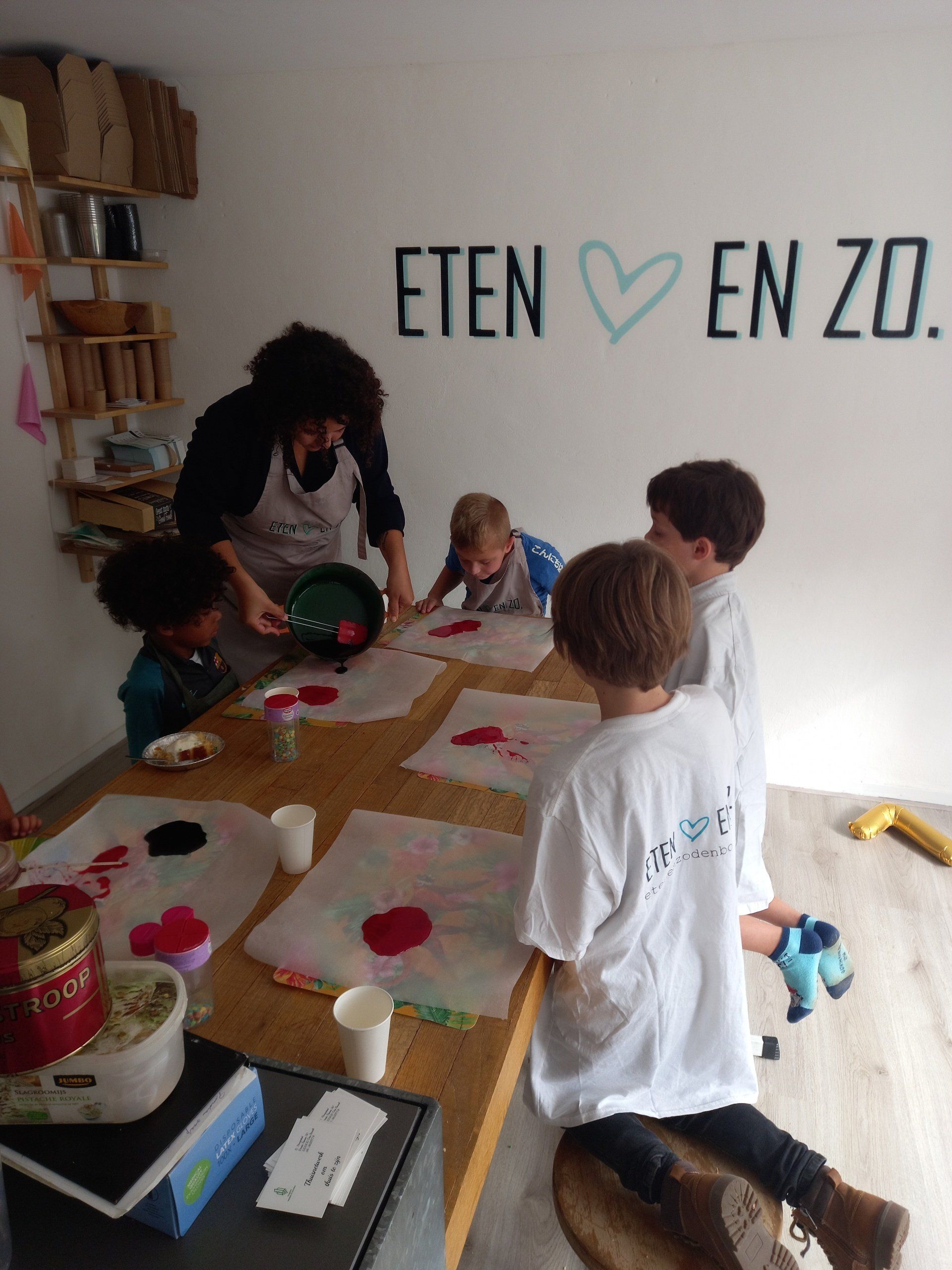 Een volwassene leert een groep kinderen knutselen met rode materialen aan een tafel in een kamer met het opschrift 