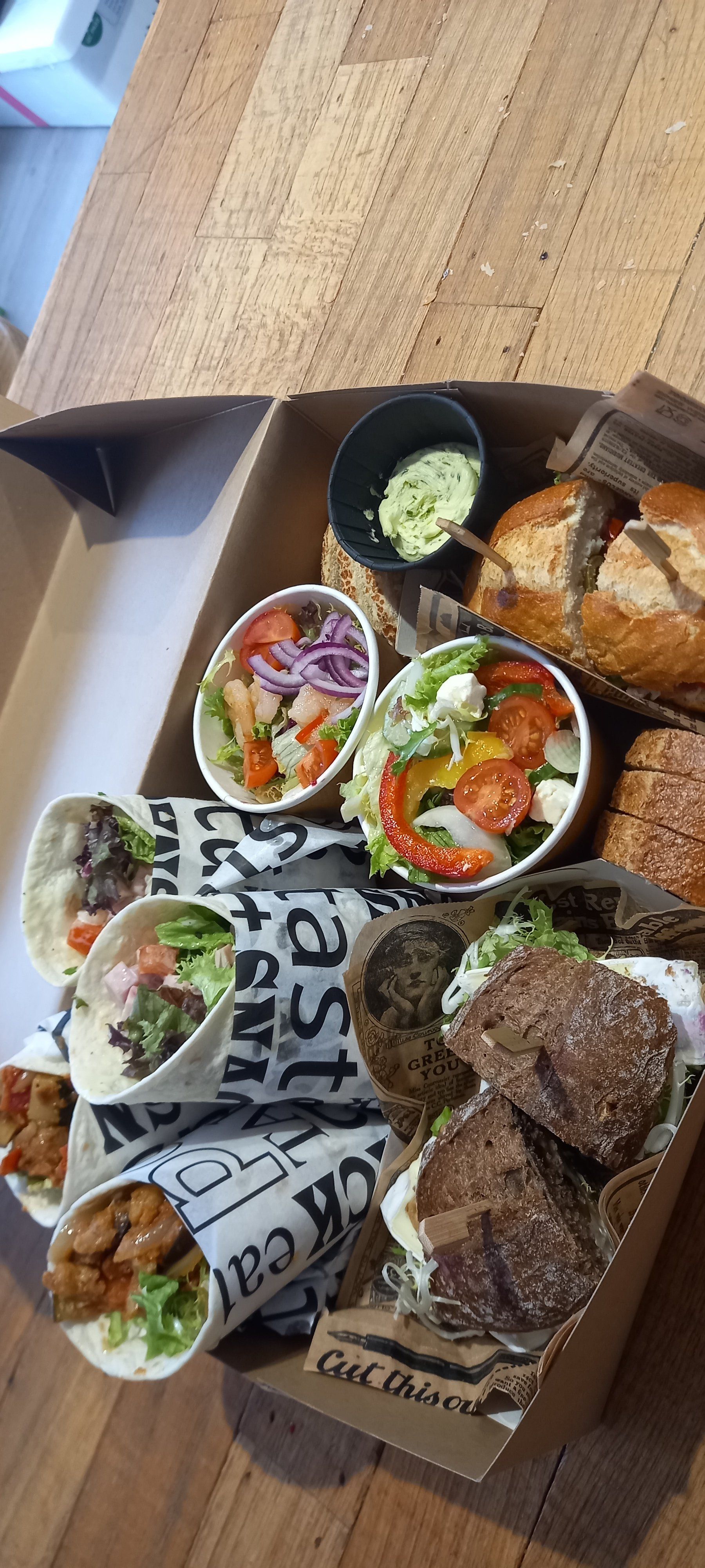 Een kartonnen doos gevuld met wraps, twee kleine schaaltjes salade, een bakje groene dipsaus en geroosterd brood op een houten tafel.